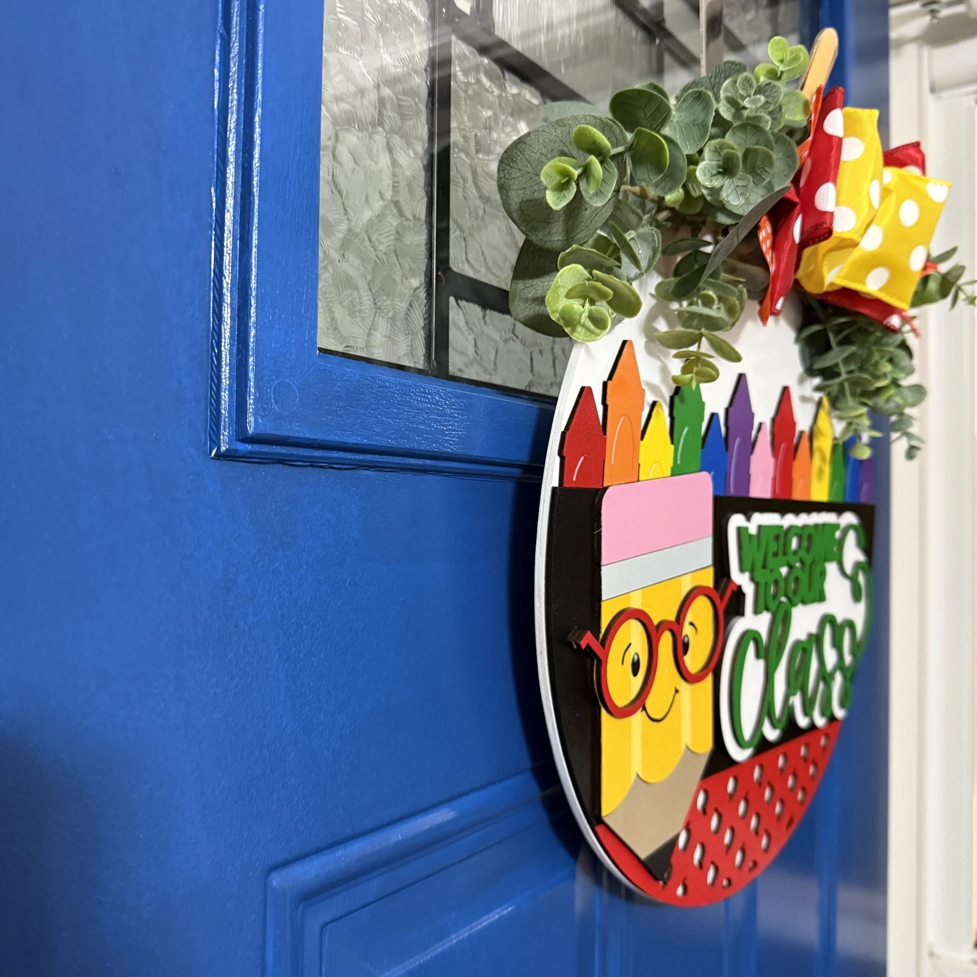 Side view of “Welcome to Our Class” door hanger featuring smiling pencil, rainbow crayon row, and red/yellow polka dot bow with greenery on a dark blue door.