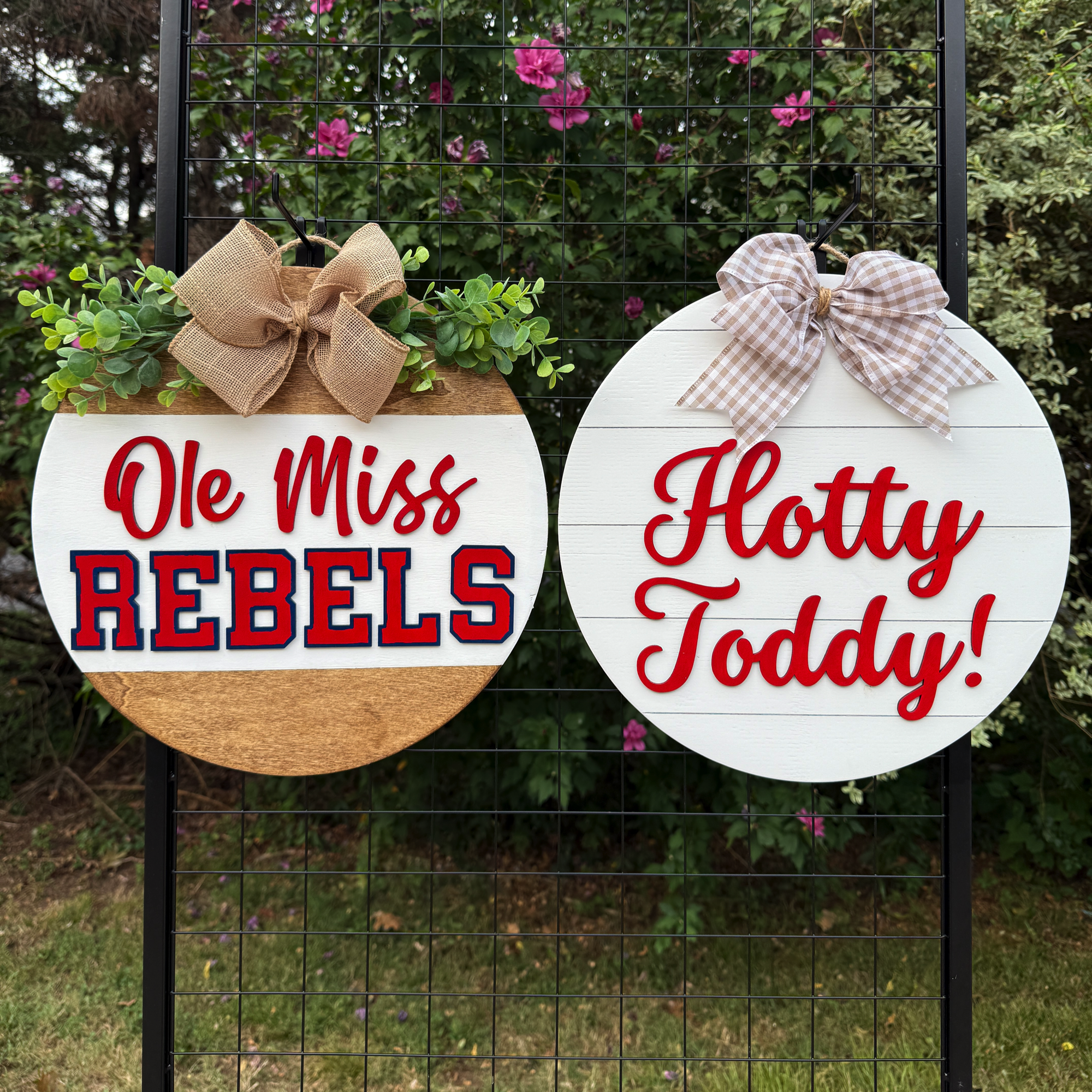 Two round Ole Miss-themed door hangers displayed outdoors on a black grid with greenery in the background; one reads “Ole Miss Rebels” and the other “Hotty Toddy!”