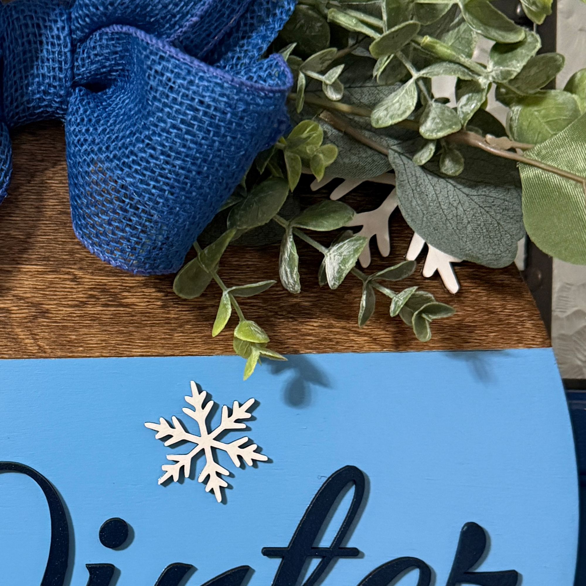 Close up of Winter Is Stupid text and layered white snowflake accents on blue painted wooden door hanger