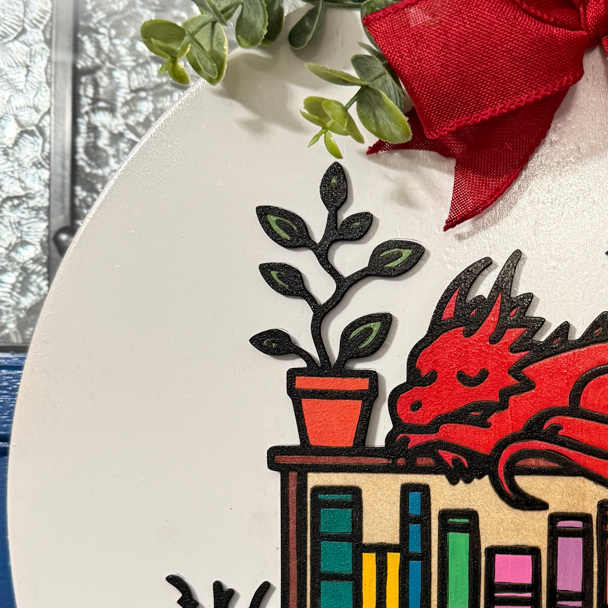 Close-up of top section with red bow, leafy greenery, and small cat figure on bookshelf