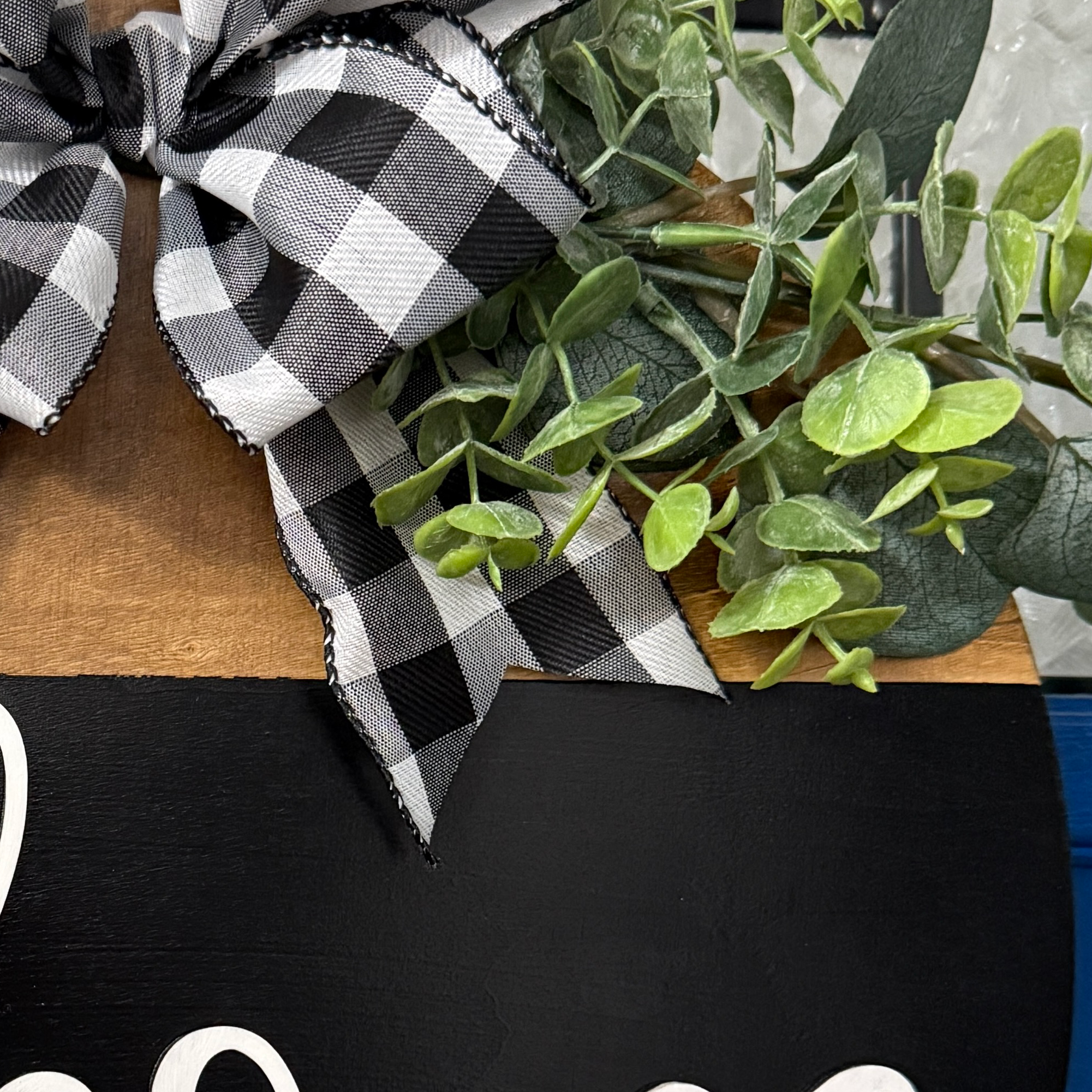 Close up of Welcome door hanger on stained base with black background, faux with greenery and coordinating bow