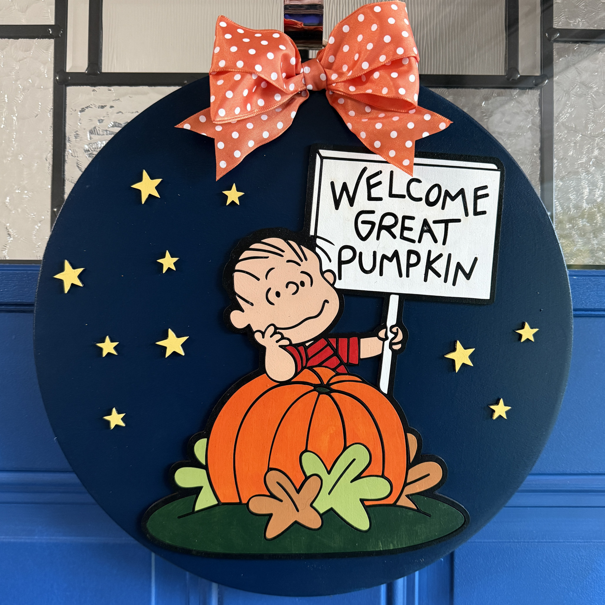 Front view of Peanuts door hanger featuring Linus holding a “Welcome Great Pumpkin” sign, sitting in a pumpkin patch with stars on a navy background
