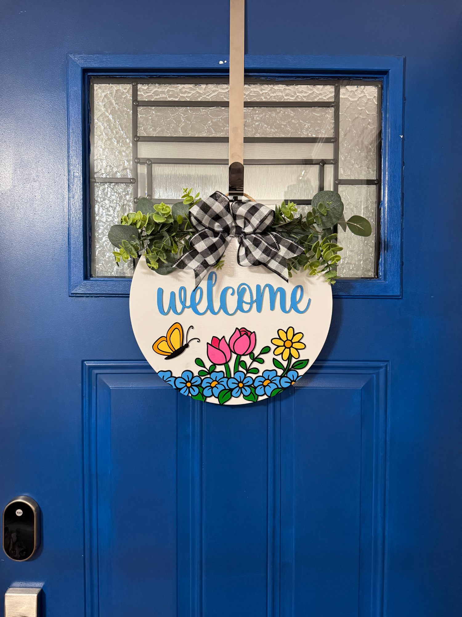 Full view of Welcome spring door hanger displayed on bright blue front door with buffalo check bow and vibrant floral garden design