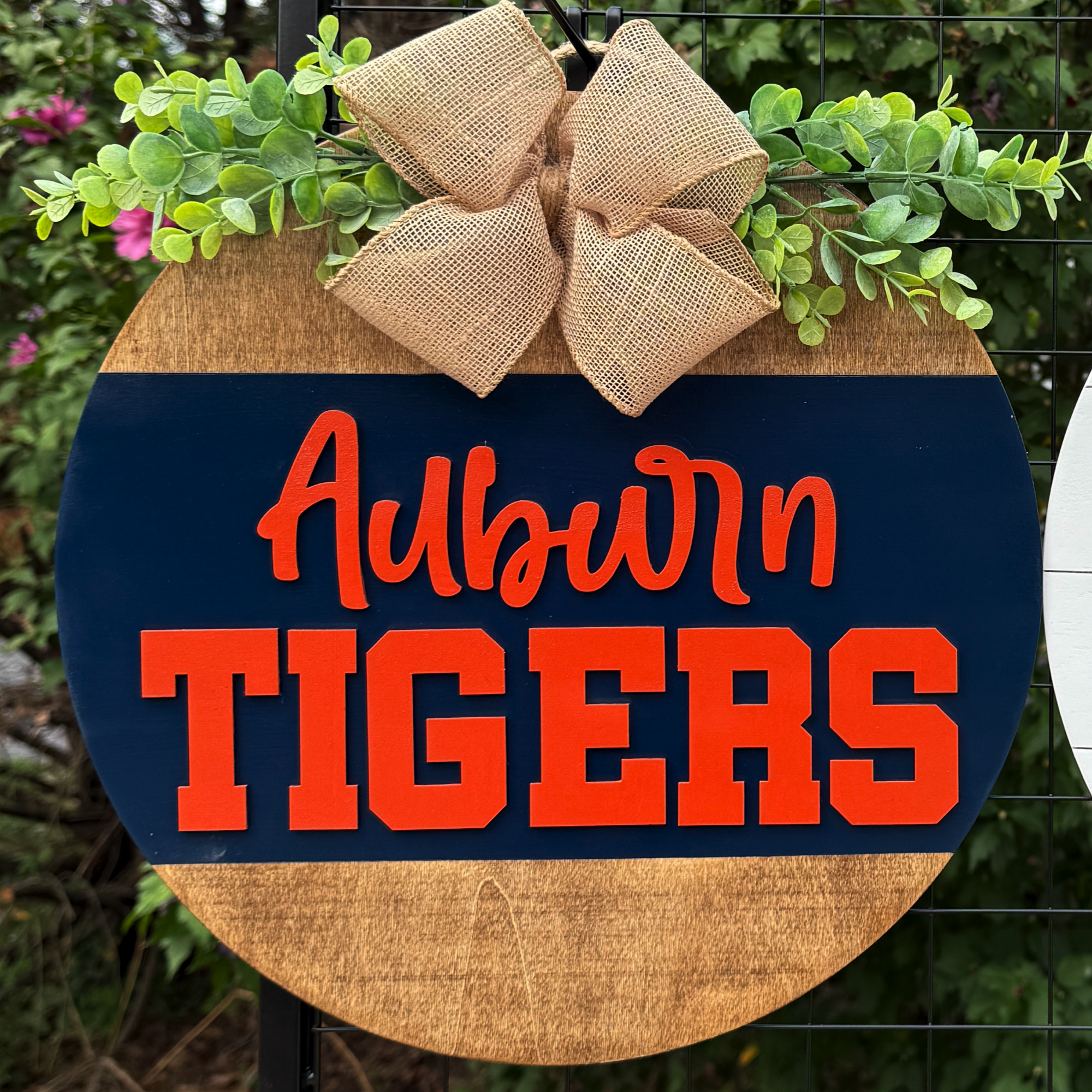 Round Auburn Tigers door hanger with navy and orange lettering, greenery accents, and burlap bow on rustic wood background