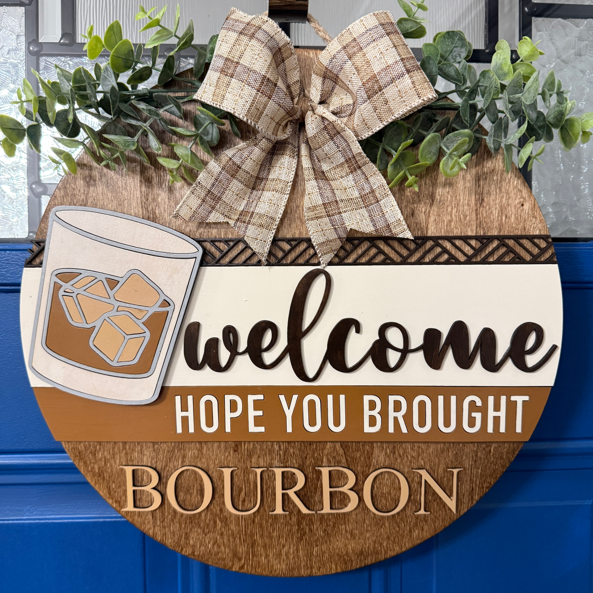 Front view of round wooden bourbon welcome door hanger with whiskey glass, greenery, and “Hope you brought bourbon” text
