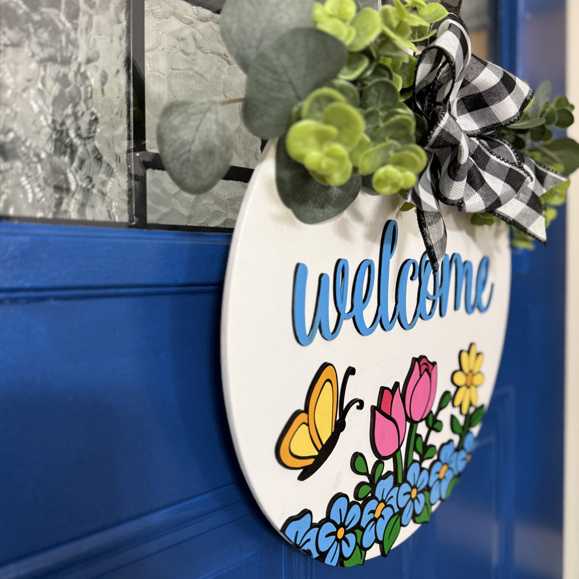 Angled side view of Welcome floral wood door hanger showing layered thickness, greenery, and dimensional blue script lettering