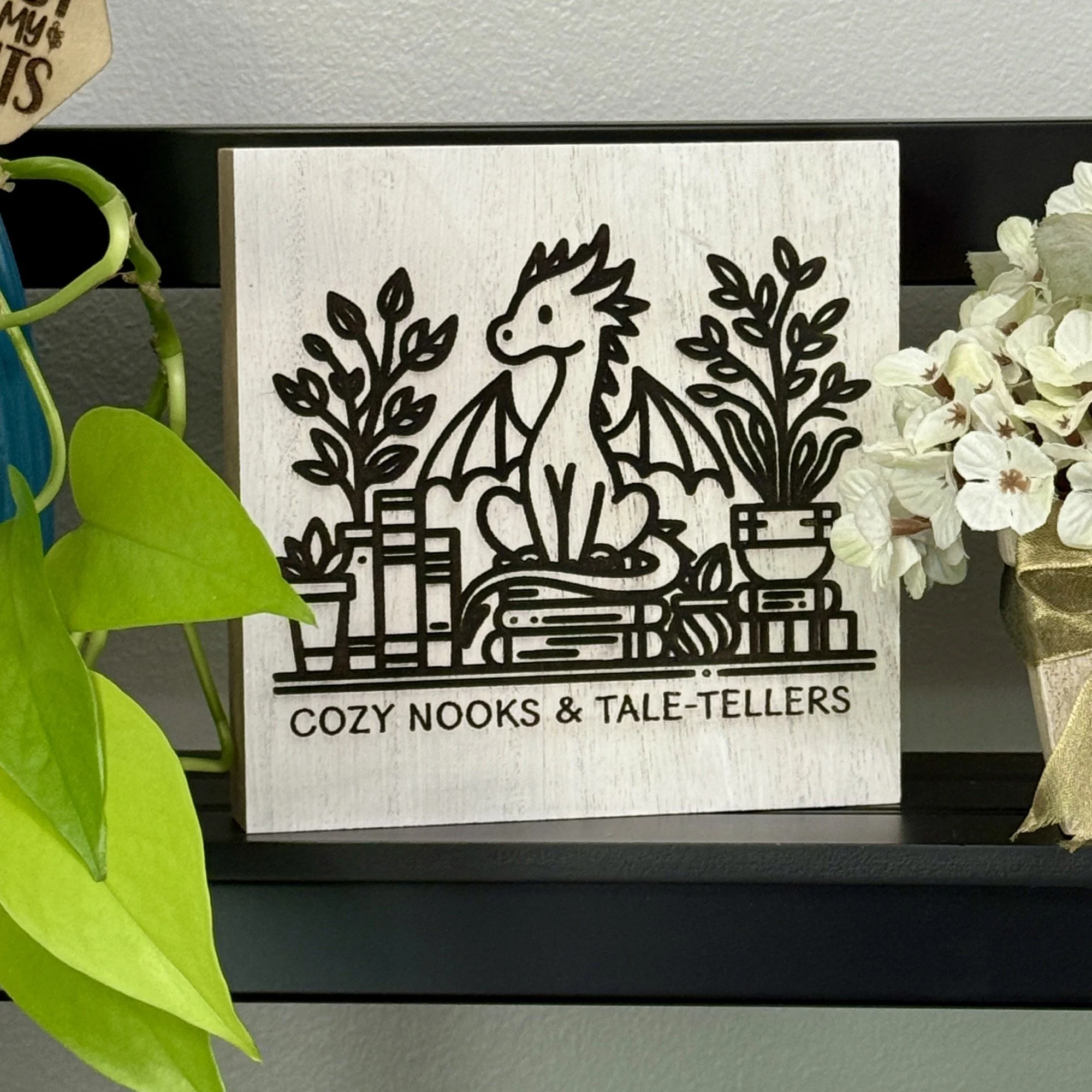 White-washed wood shelf sitter engraved with a seated dragon among stacked books and potted plants, displayed on a black shelf with greenery and flowers.