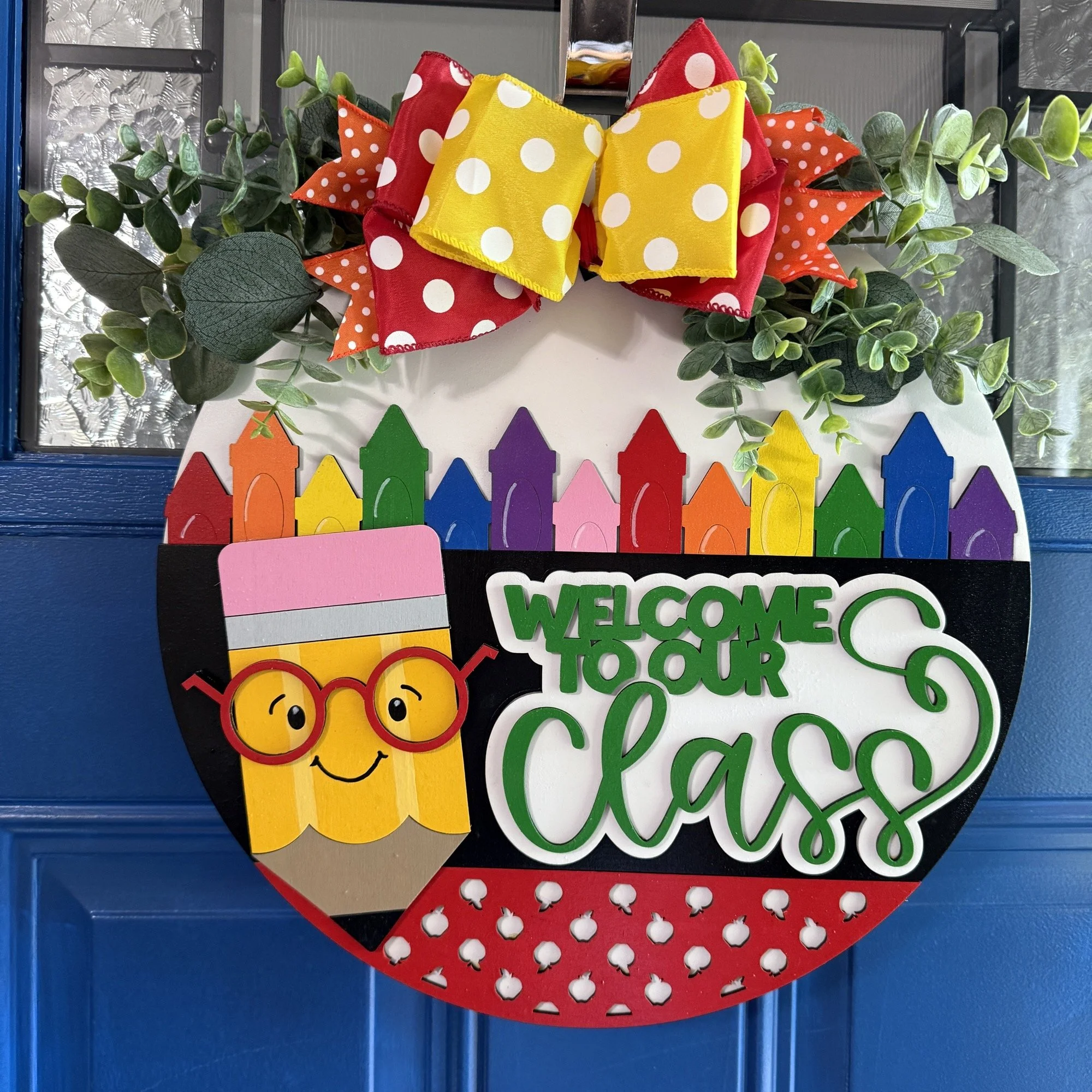 Front view of “Welcome to Our Class” door hanger featuring smiling pencil, rainbow crayon row, and red/yellow polka dot bow with greenery on a dark blue door