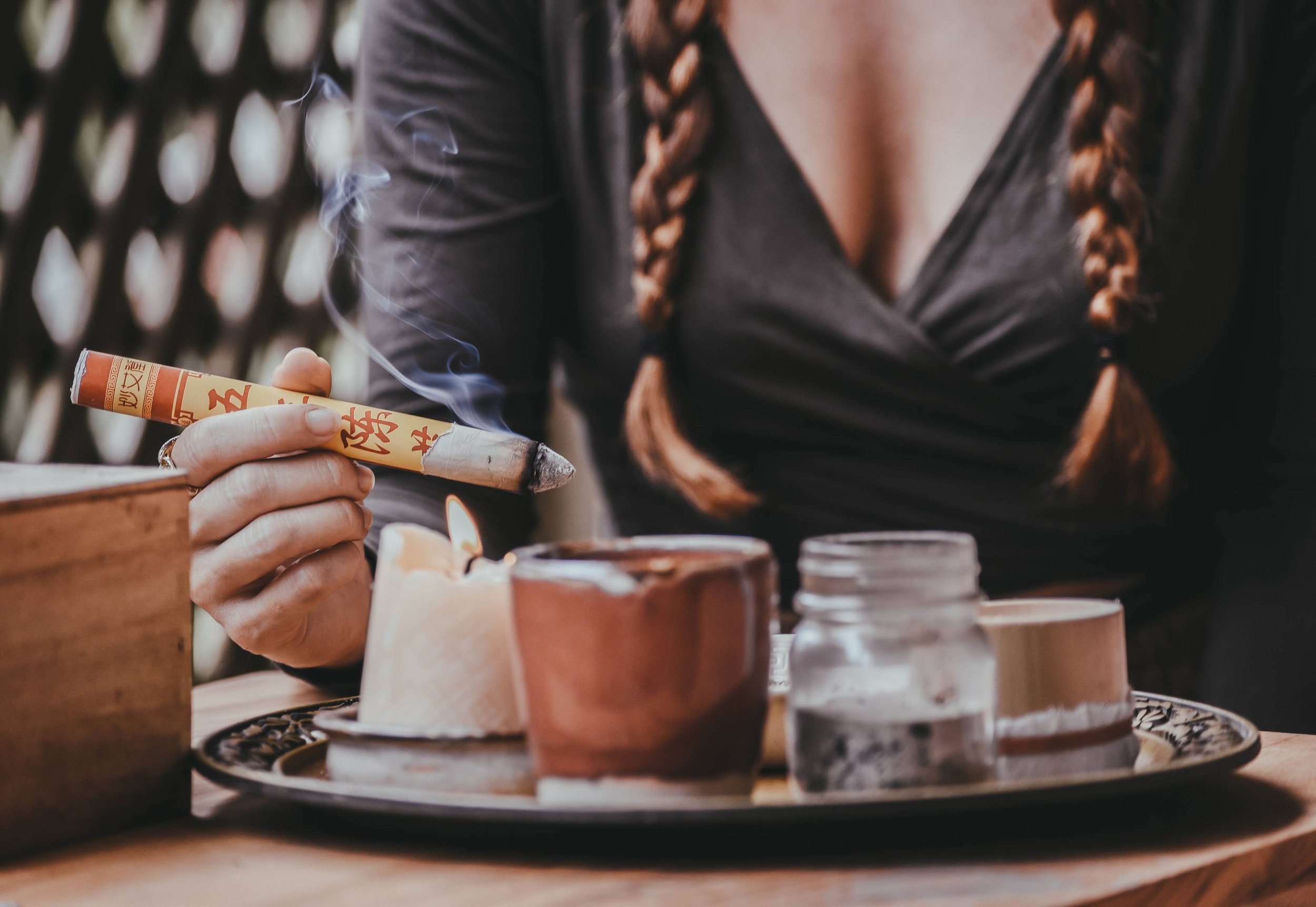 Person holding a smoldering incense stick near candles on a table, with braided hair visible.