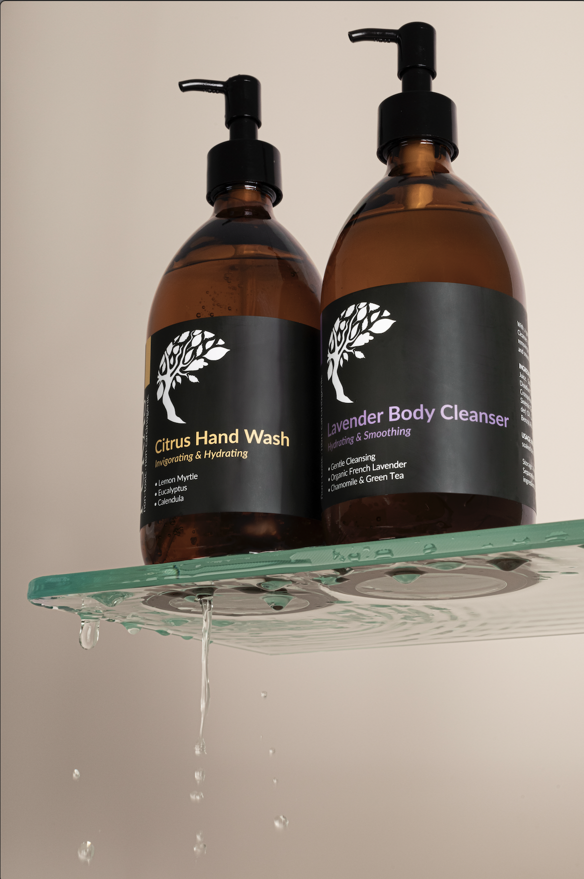 Two brown bottles with black pump dispensers on a glass shelf, one labeled Citrus Hand Wash, the other Lavender Body Cleanser, with water droplets on the shelf.