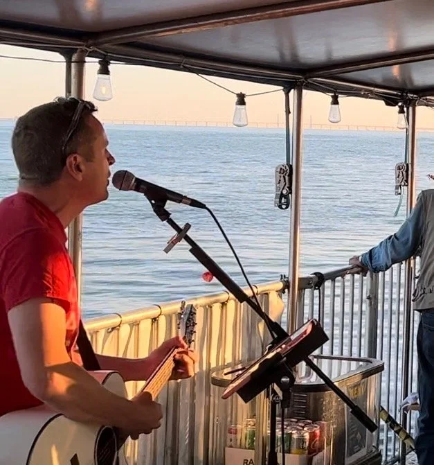 Man playing guitar and singing into a microphone on a boat with water view in the background.LiveFlotten Malmö. Malmö City. Summer in Malmö. Flotten Malmö. Malmösommar. Vår i Malmö Spring Malmö