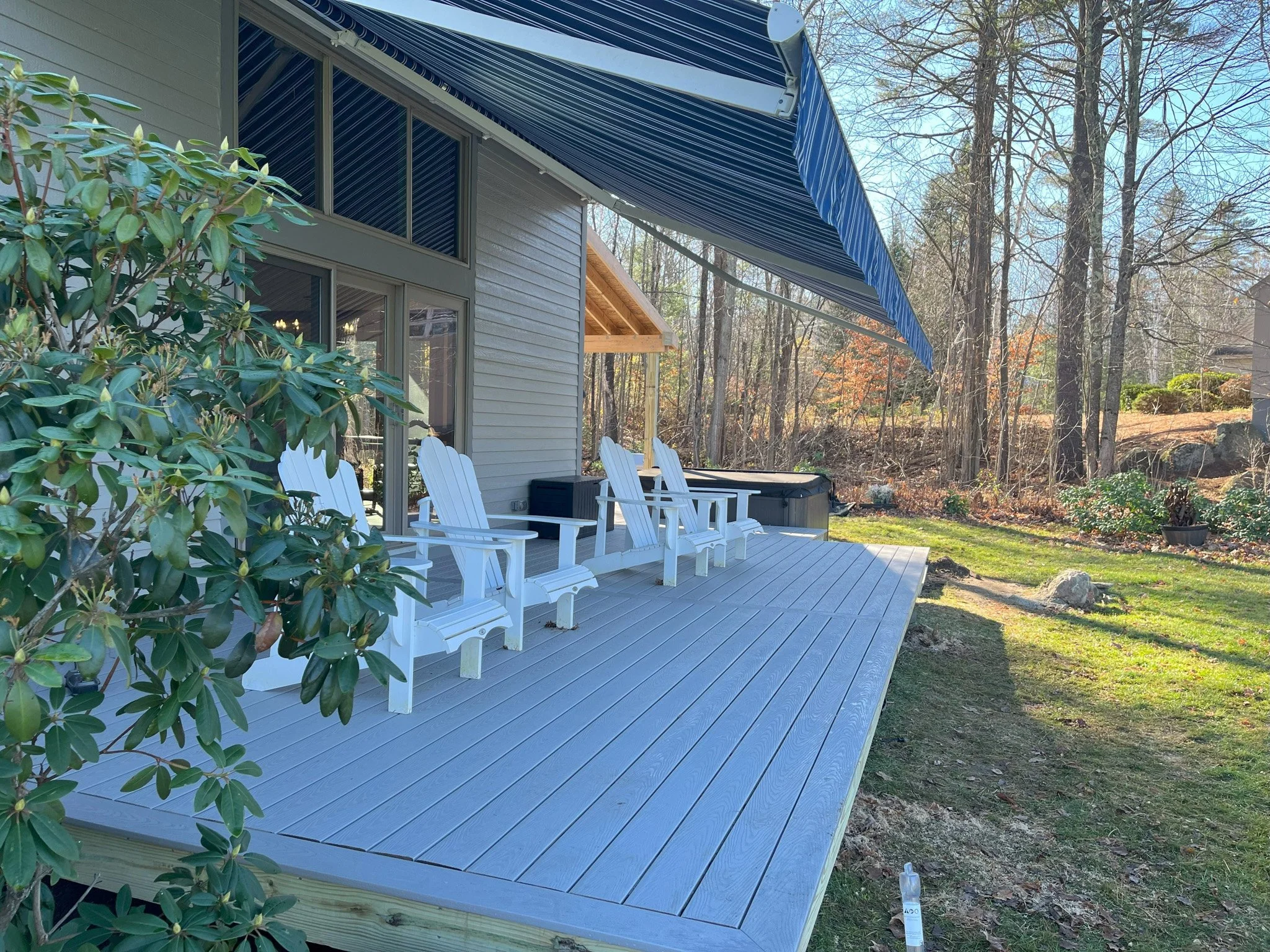 A backyard deck with three white Adirondack chairs and a hot tub, shaded by a blue and white striped awning, with trees and bushes in the background.