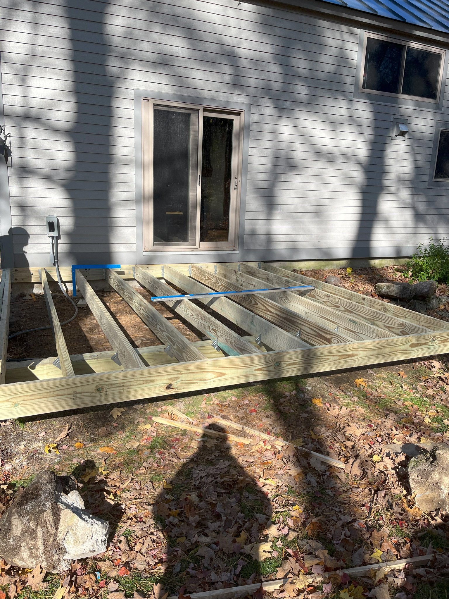 Construction of a wooden deck in progress outside a house with gray siding, sliding door, and windows, with shadows of trees and a person taking the photo visible on the ground covered with fallen leaves.
