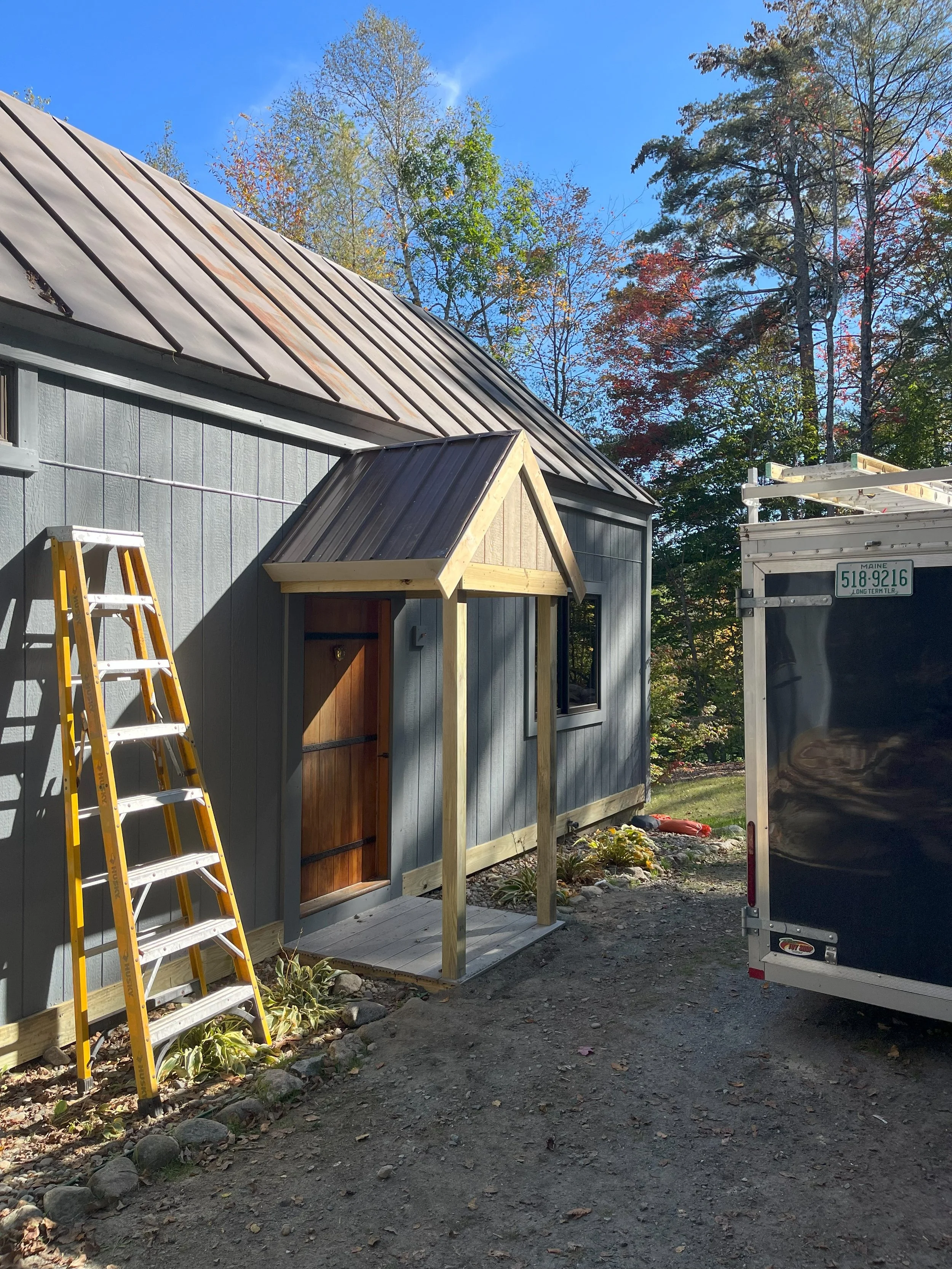 Construction of a small porch with a gabled roof on a house with gray wooden siding, next to a ladder and a trailer, surrounded by trees with fall foliage.