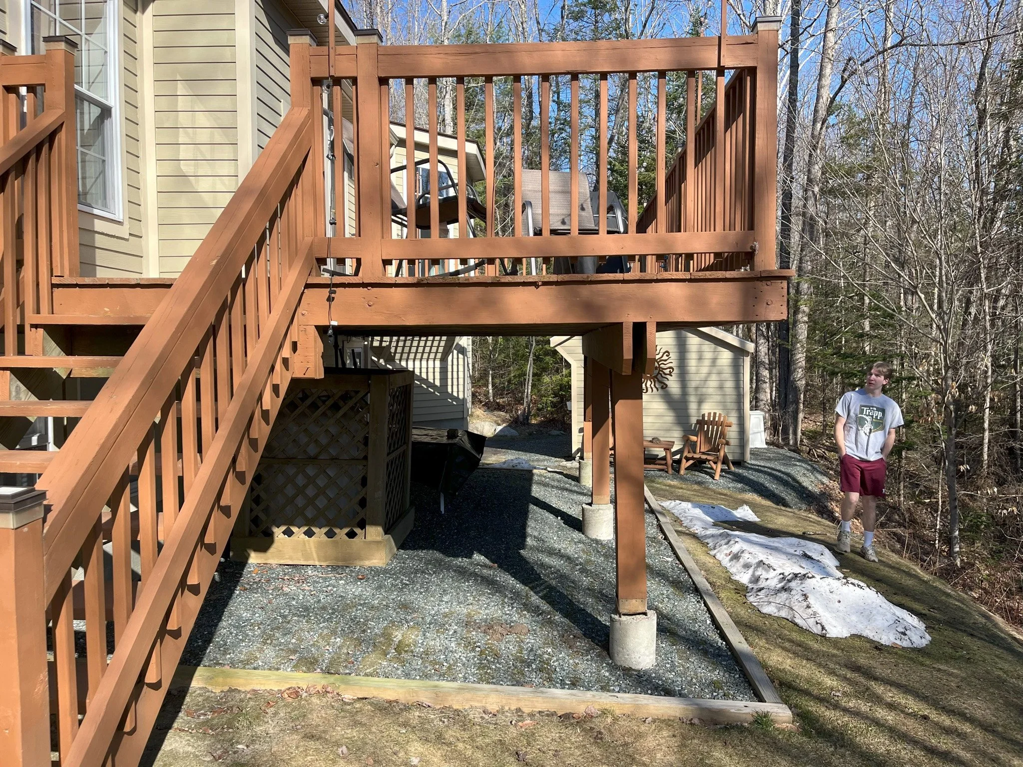 A backyard wooden deck with stairs and a man standing on the ground nearby, surrounded by trees and some patches of snow on the ground.