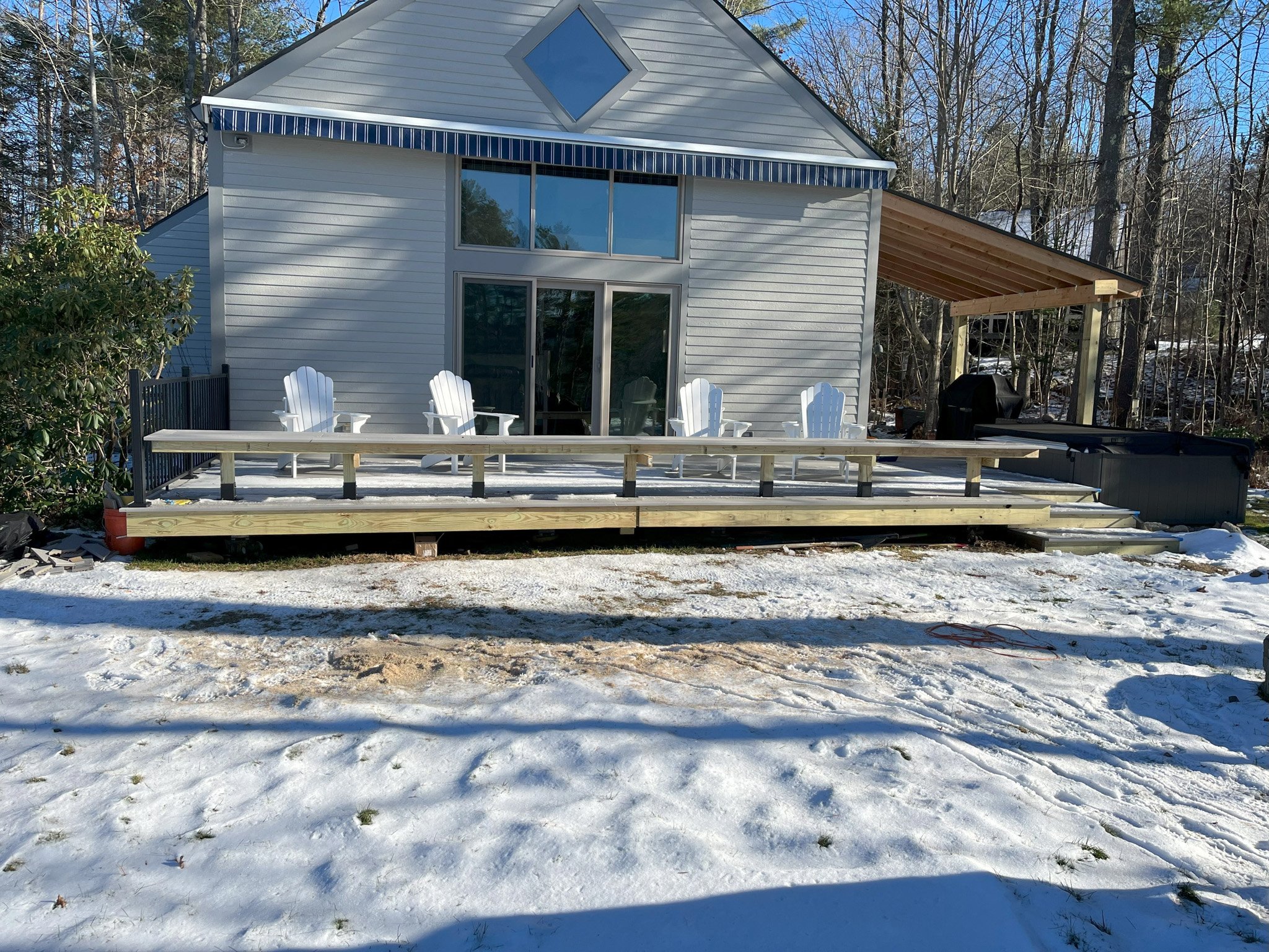 A house with a new wooden deck under construction, featuring four white Adirondack chairs, and a covered area with a grill, set in a snowy backyard surrounded by trees.