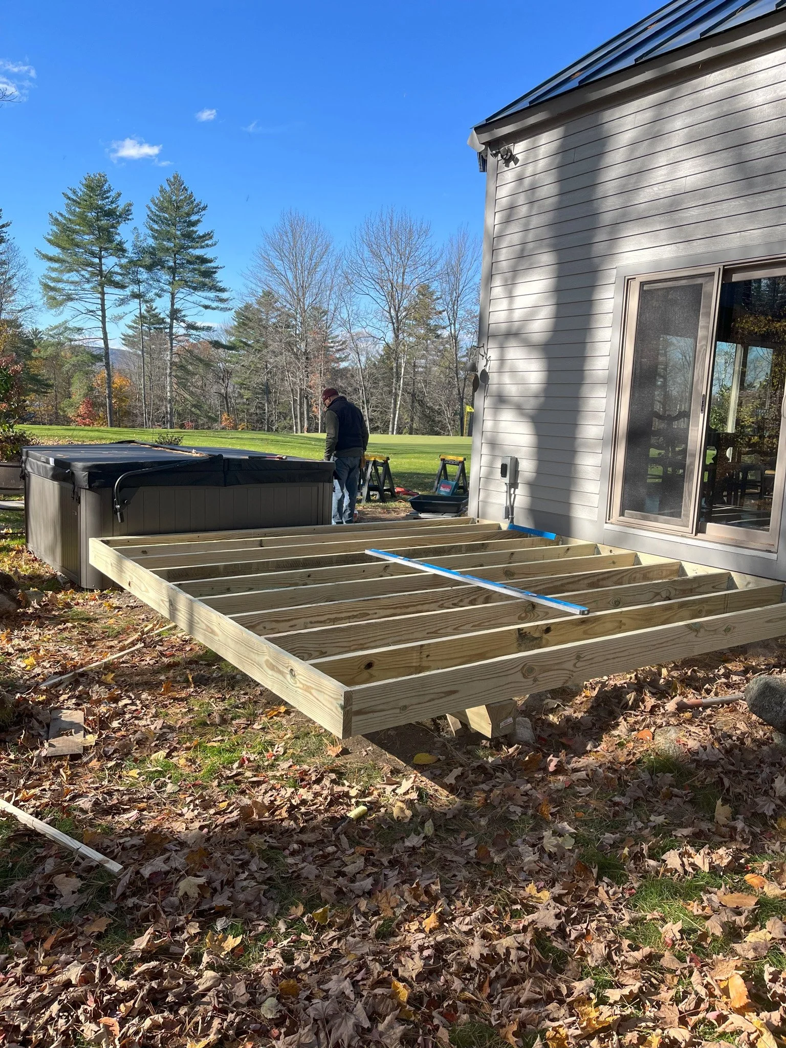 Wooden deck frame being built outside a house, with a person working nearby during autumnal weather.