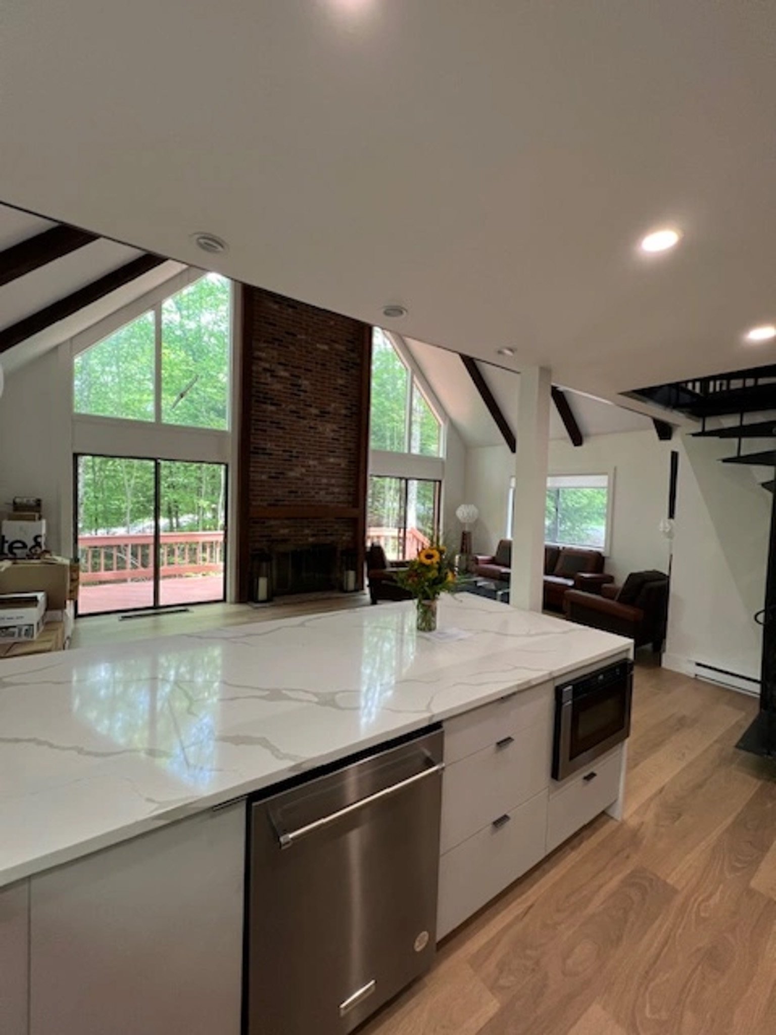 Modern kitchen with white countertop, stainless steel appliances, and open view of living area with large windows, brick fireplace, and cozy seating.