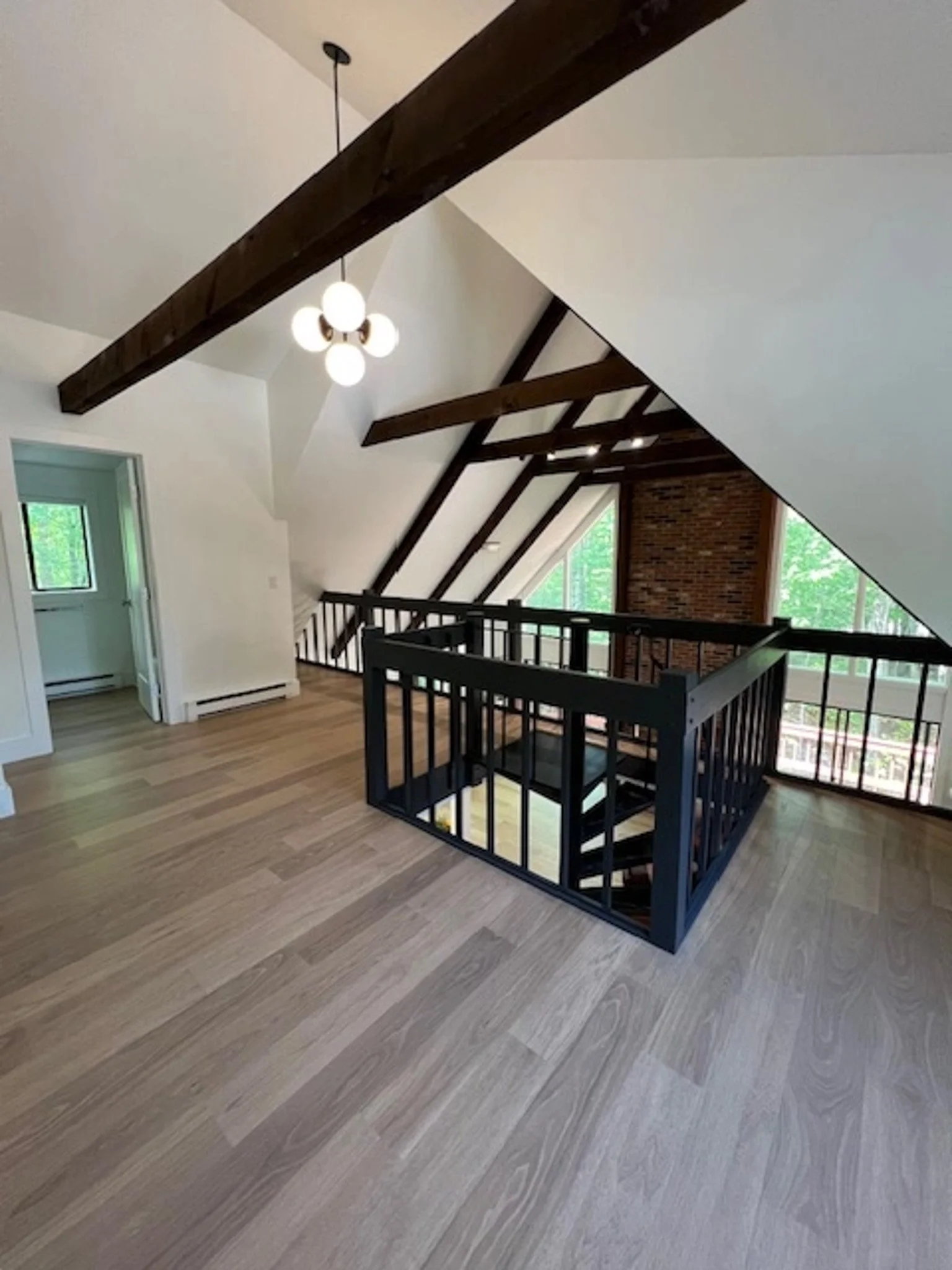 A spacious attic with sloped ceilings, wooden beams, a black railing around the staircase, large windows, and a modern light fixture hanging from the ceiling.