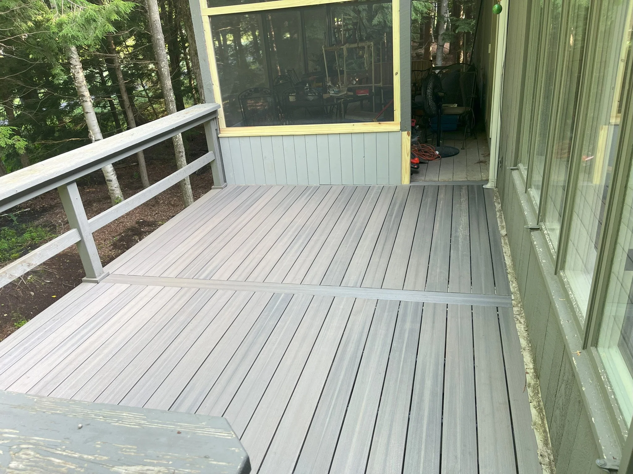 A new grey wooden deck attached to a house, overlooking a wooded area with trees. The deck has a simple railing on one side and is adjacent to a screened porch with a glass door leading inside.
