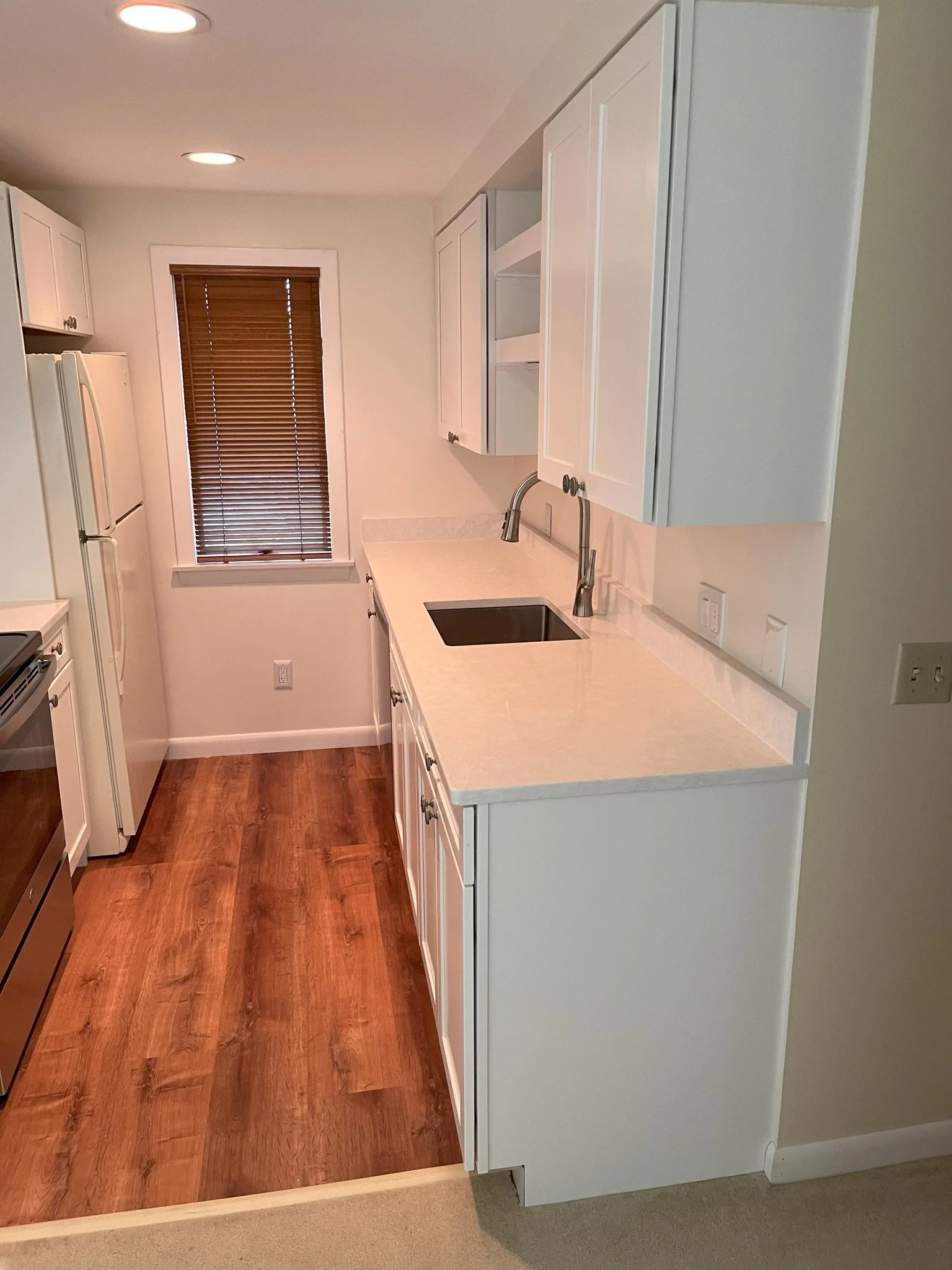 A small kitchen with white cabinets, a white countertop, and a single sink. There is a window with brown blinds and a wooden floor. The kitchen appliances include a white refrigerator and a white oven/stove.
