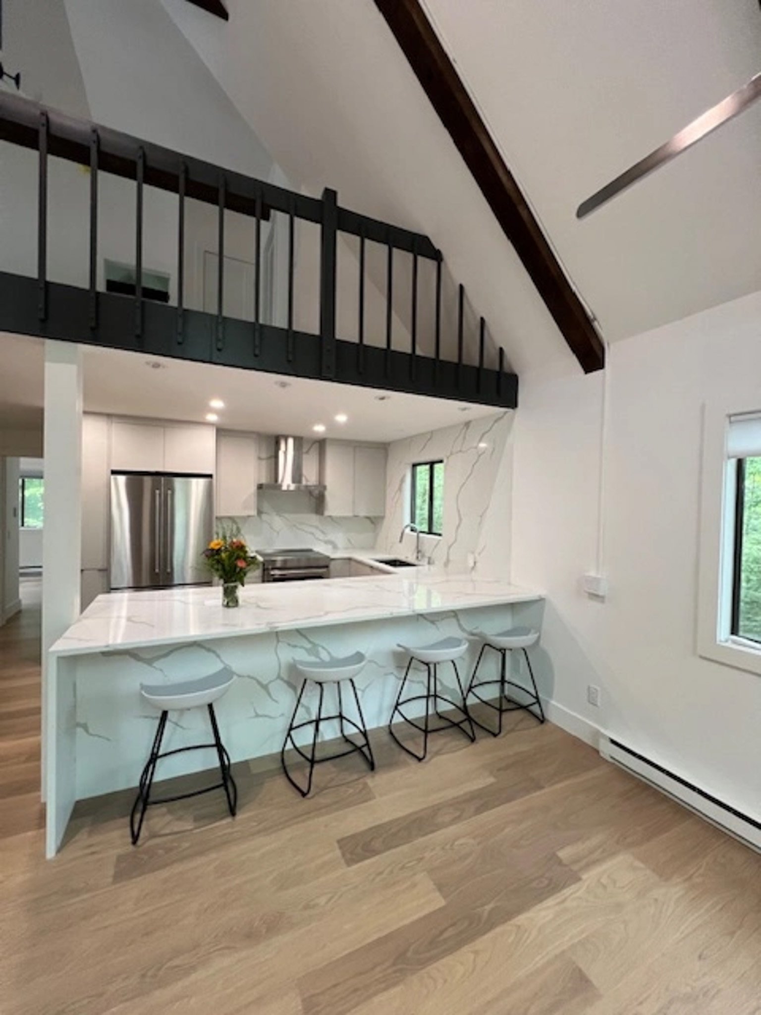 Modern kitchen with white marble island, four white barstools with black legs, stainless steel appliances, and hardwood floors.