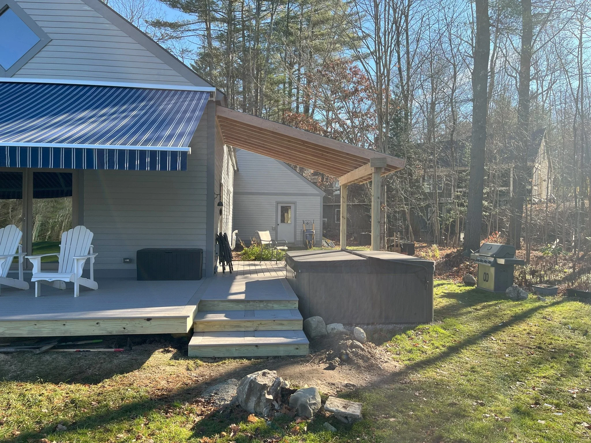 Backyard with a wooden deck, white chairs, a blue striped awning, a grill, and a wooded area in the background.