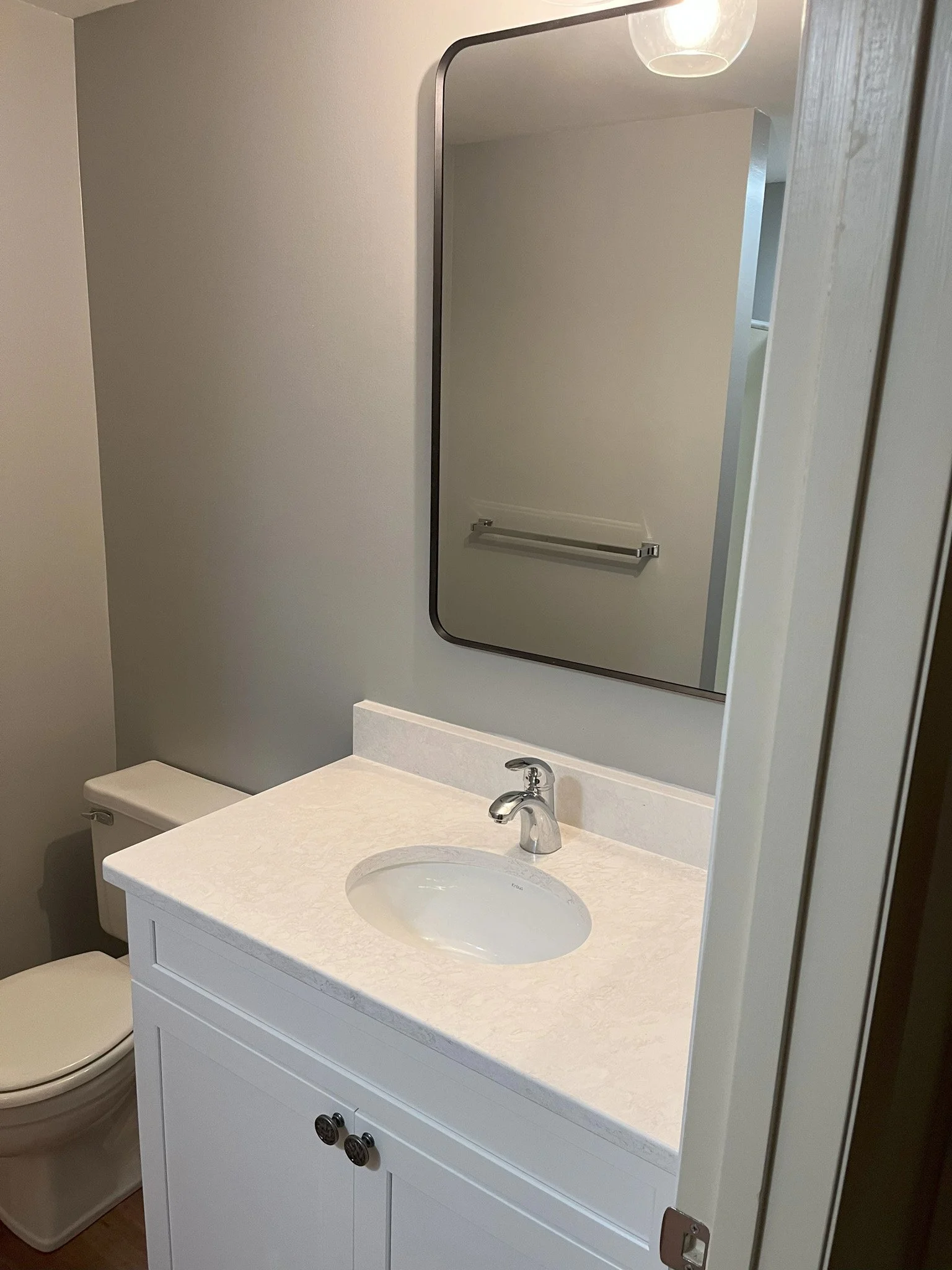 A small bathroom with a white vanity sink, a mirror above, a toilet to the left, and a towel bar on the wall reflected in the mirror.