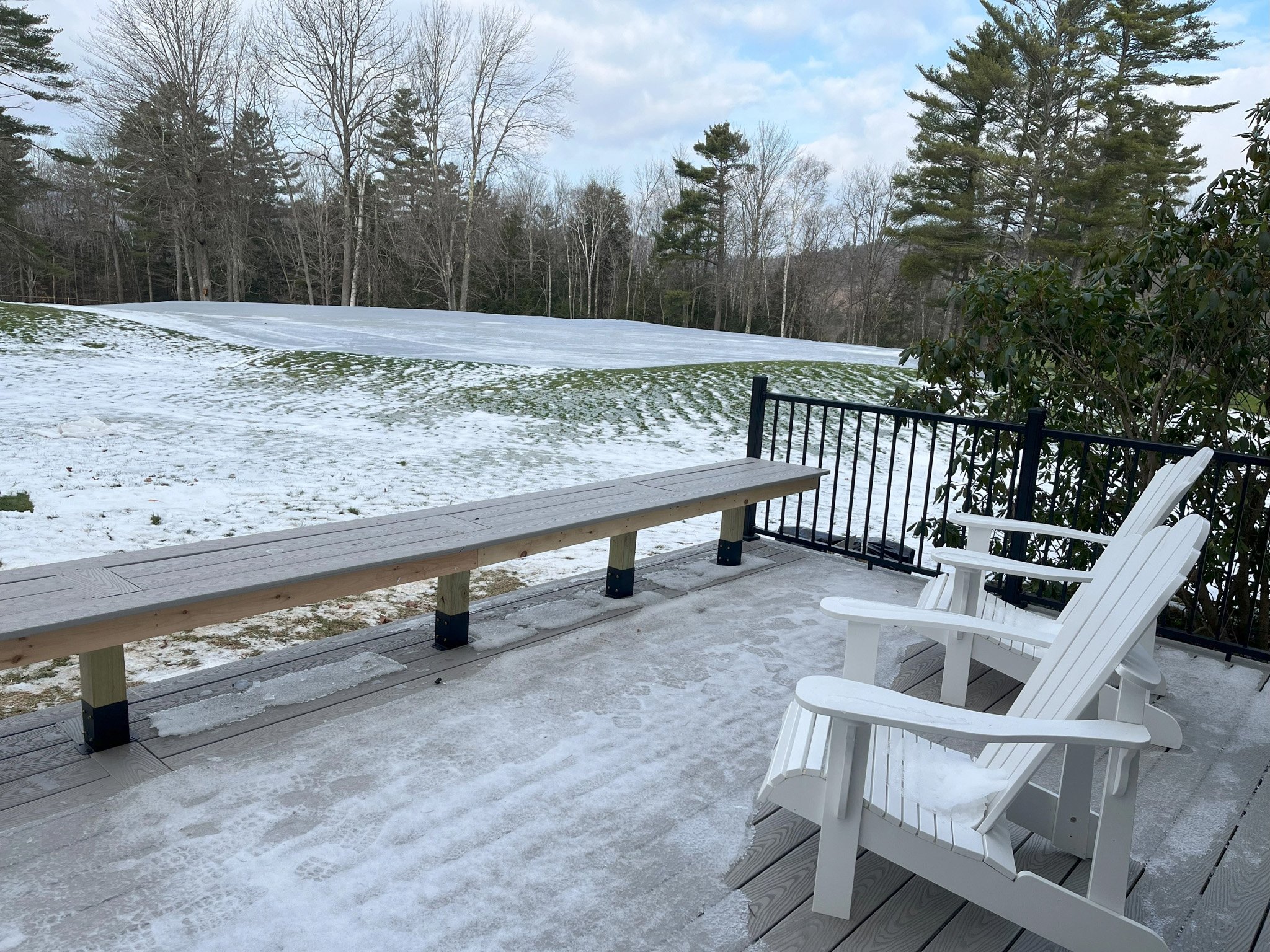A snowy deck with two white Adirondack chairs and a wooden bench, overlooking a partially snow-covered grassy yard with trees in the background.