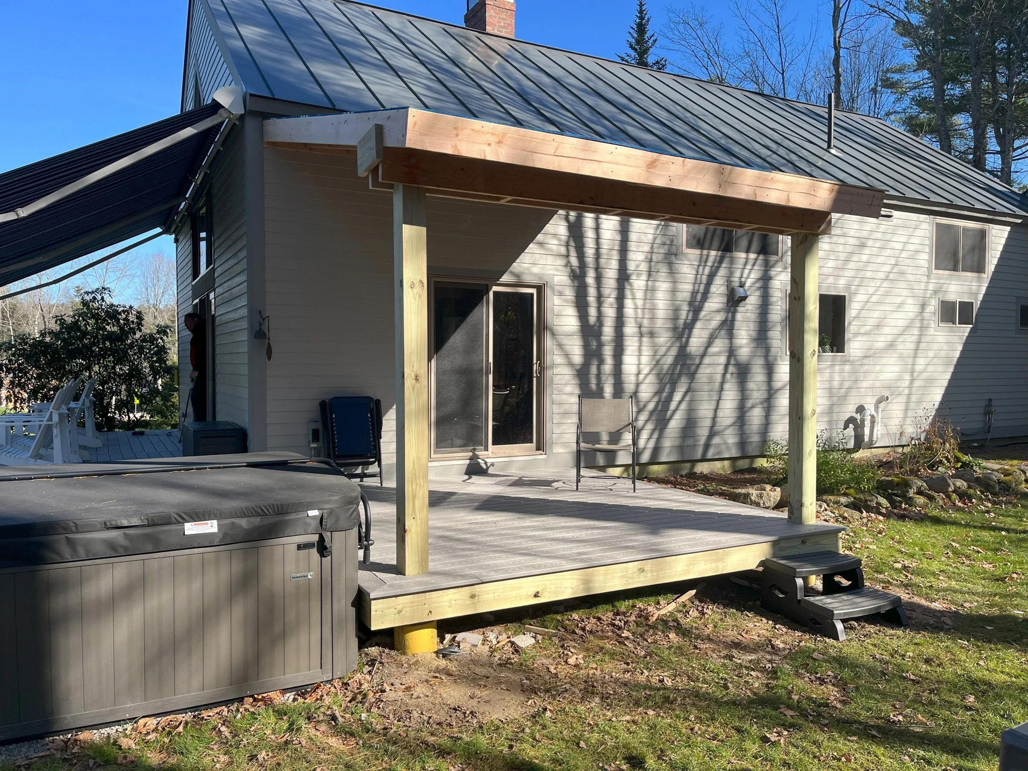 Backyard scene with a small wooden deck, sliding glass door, patio furniture, a hot tub, and a house with light-colored siding and multiple windows, with shadow of a tree cast on the house.