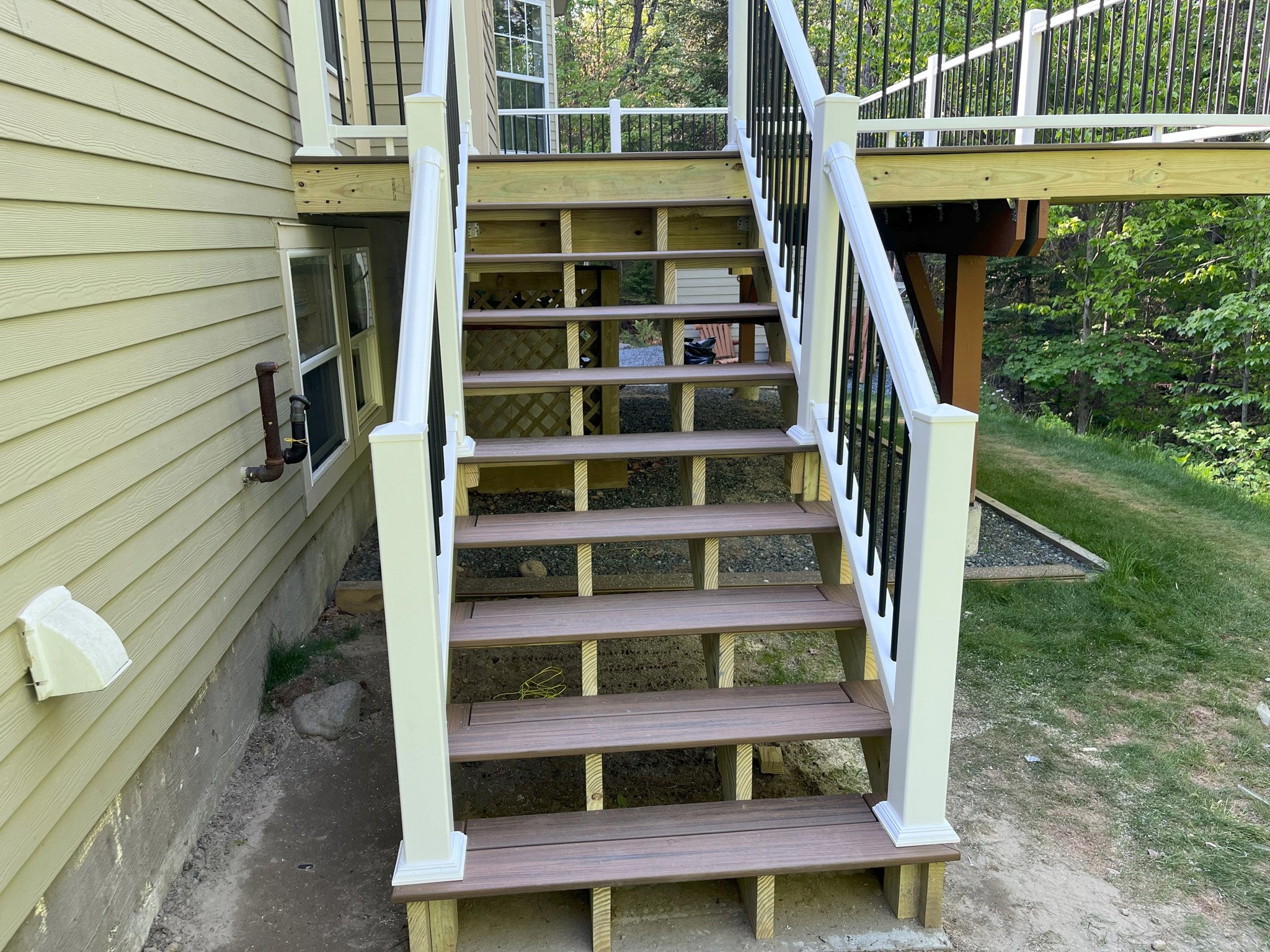 Wooden outdoor stairs with black railing leading up to a deck, attached to a yellow house with vinyl siding, in a backyard with green grass and trees.