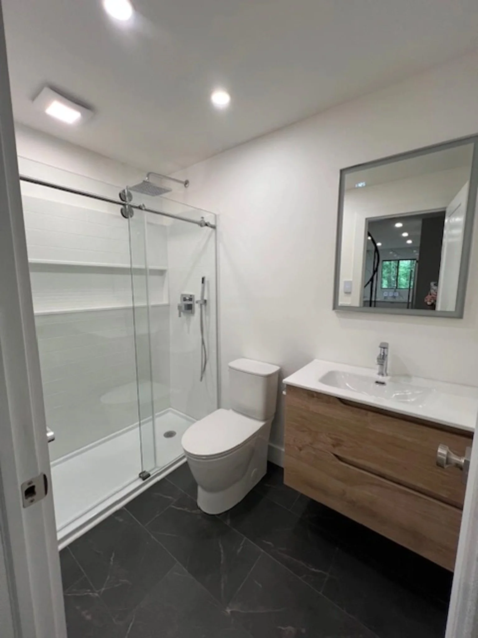 Modern bathroom with walk-in shower, white toilet, and wooden vanity with a sink, mirror, and black floor tiles.