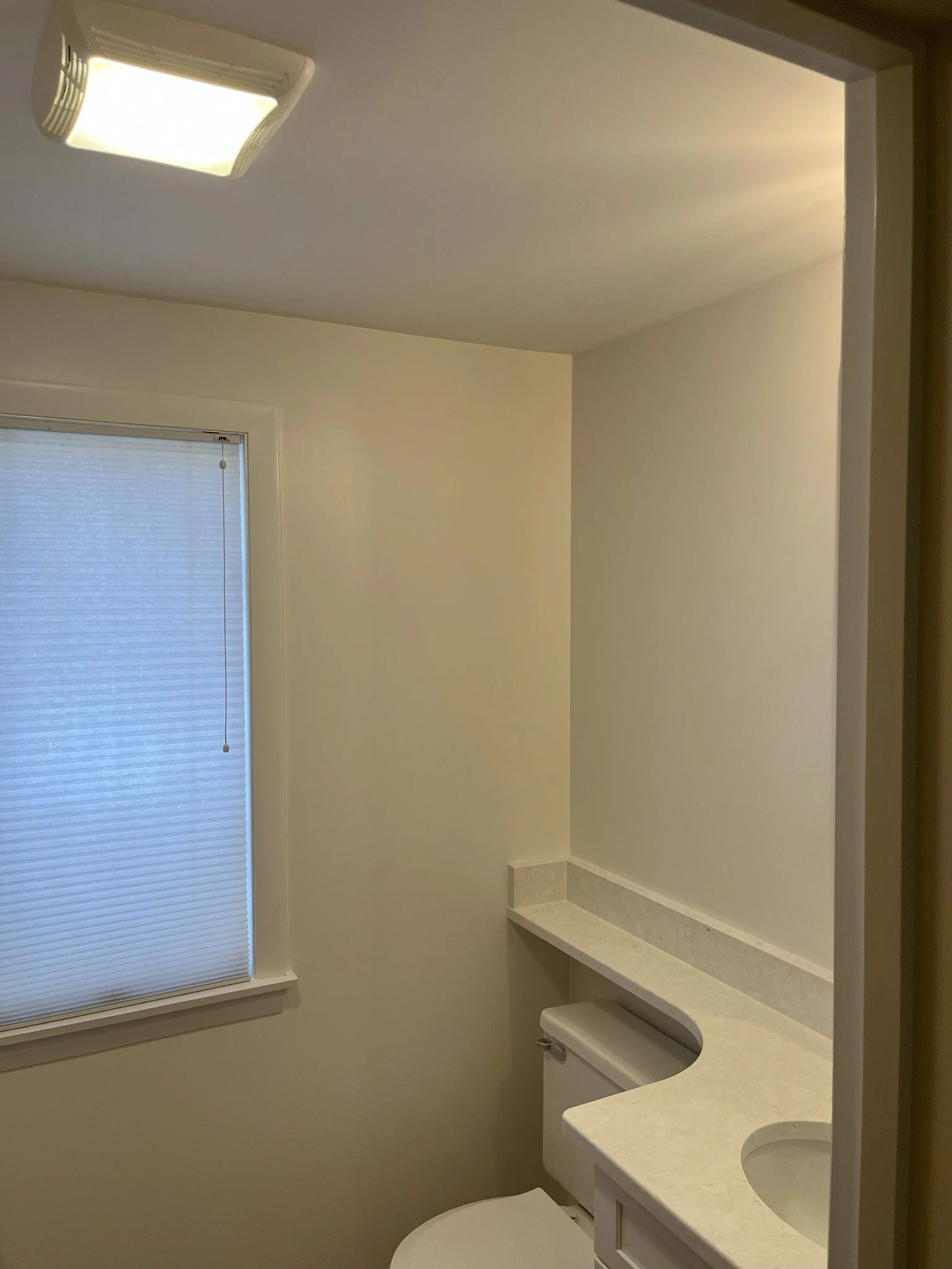 Small bathroom with beige walls, a window with closed blinds, a white toilet, and a light-colored countertop with a built-in sink.