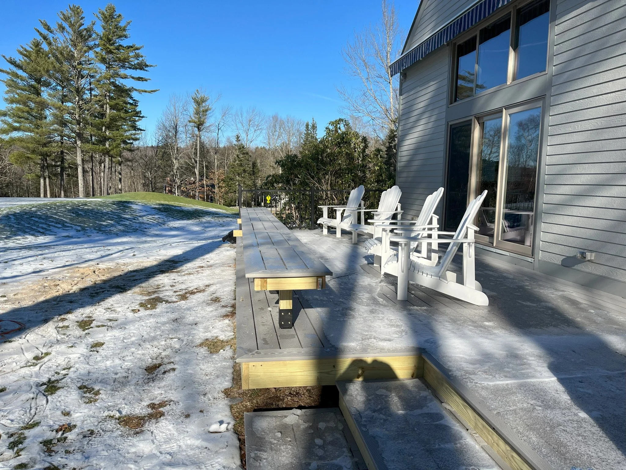 A partially constructed wooden deck attached to a house, with several white Adirondack chairs, in a snowy outdoor setting with trees in the background and a blue sky.