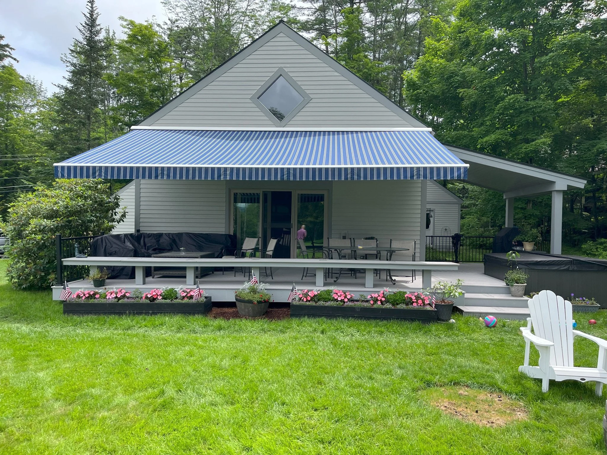 Backyard porch with blue and white striped awning, outdoor furniture, and potted plants, surrounded by green grass and trees.