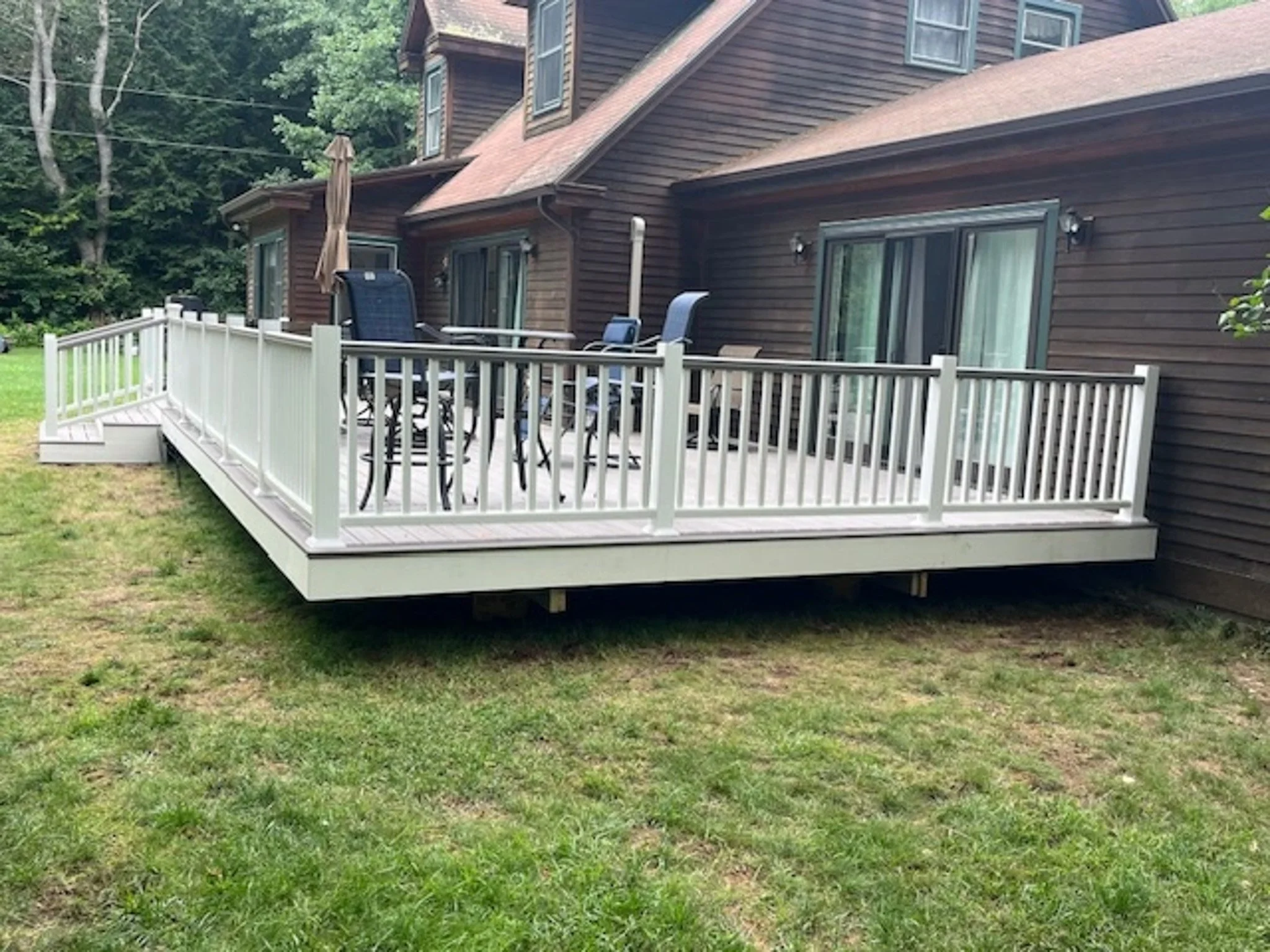 A white outdoor deck with railings attached to a brown house, featuring patio furniture and a closed umbrella, surrounded by grass and trees.