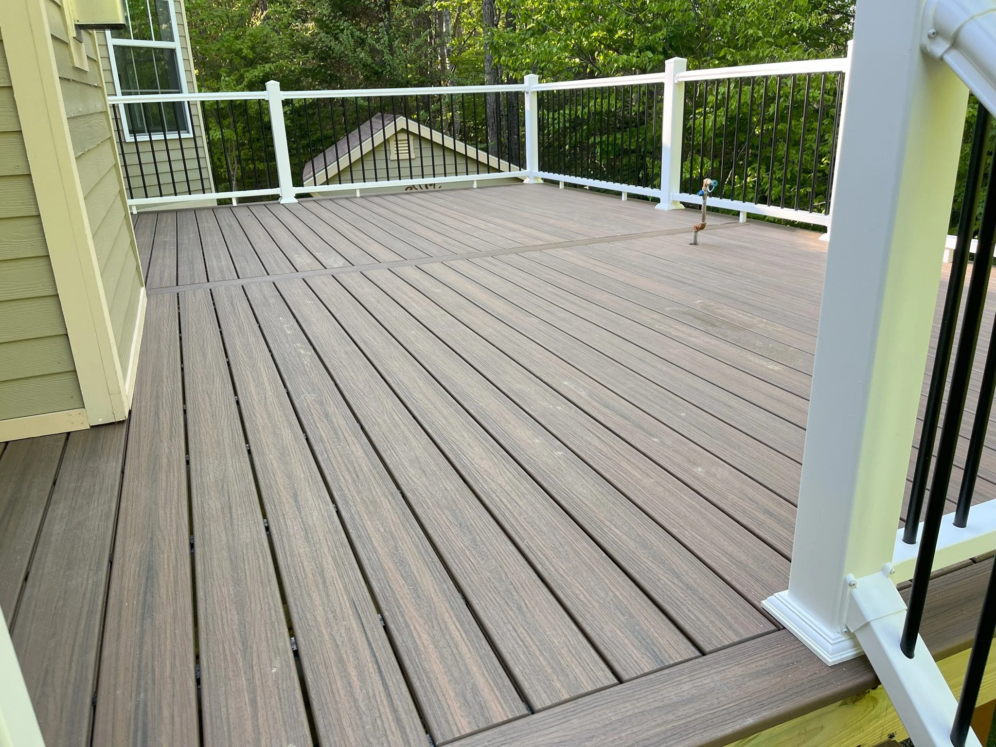 A wooden deck with a railing and a small pipe protruding from the floor, surrounded by green trees and partial view of a house.
