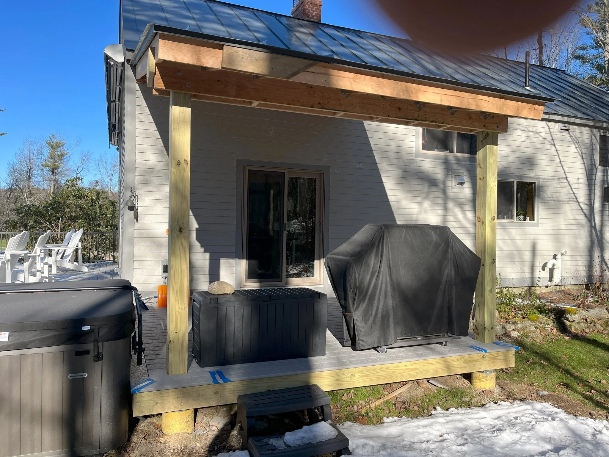 Backyard deck under construction with a wooden frame, black grill, storage box, hot tub, and outdoor furniture on the side. Snow on the ground and a house with a metal roof.