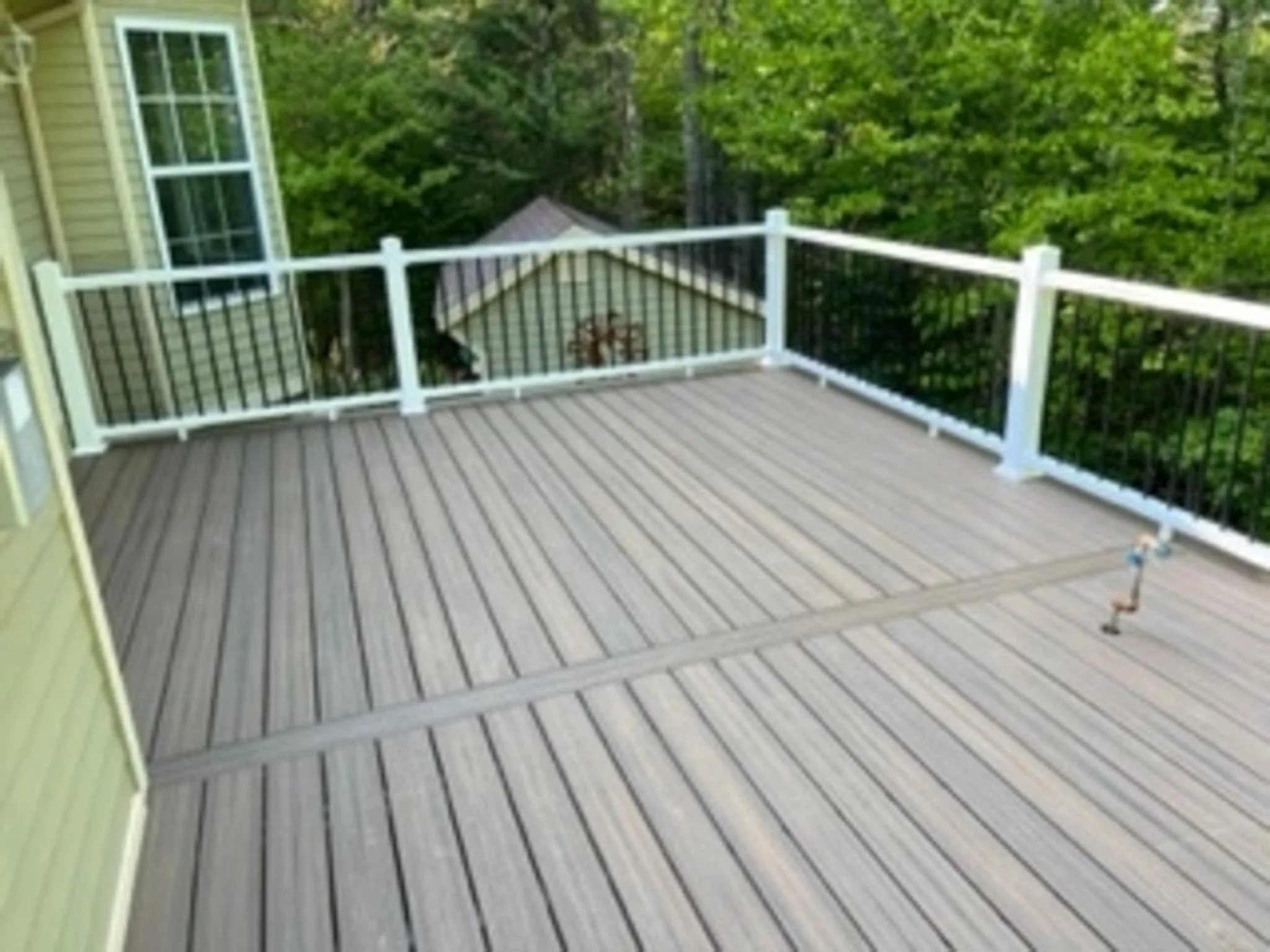 An outdoor wooden deck with a white railing, attached to a house with green siding. There are trees and a small shed or structure in the background.