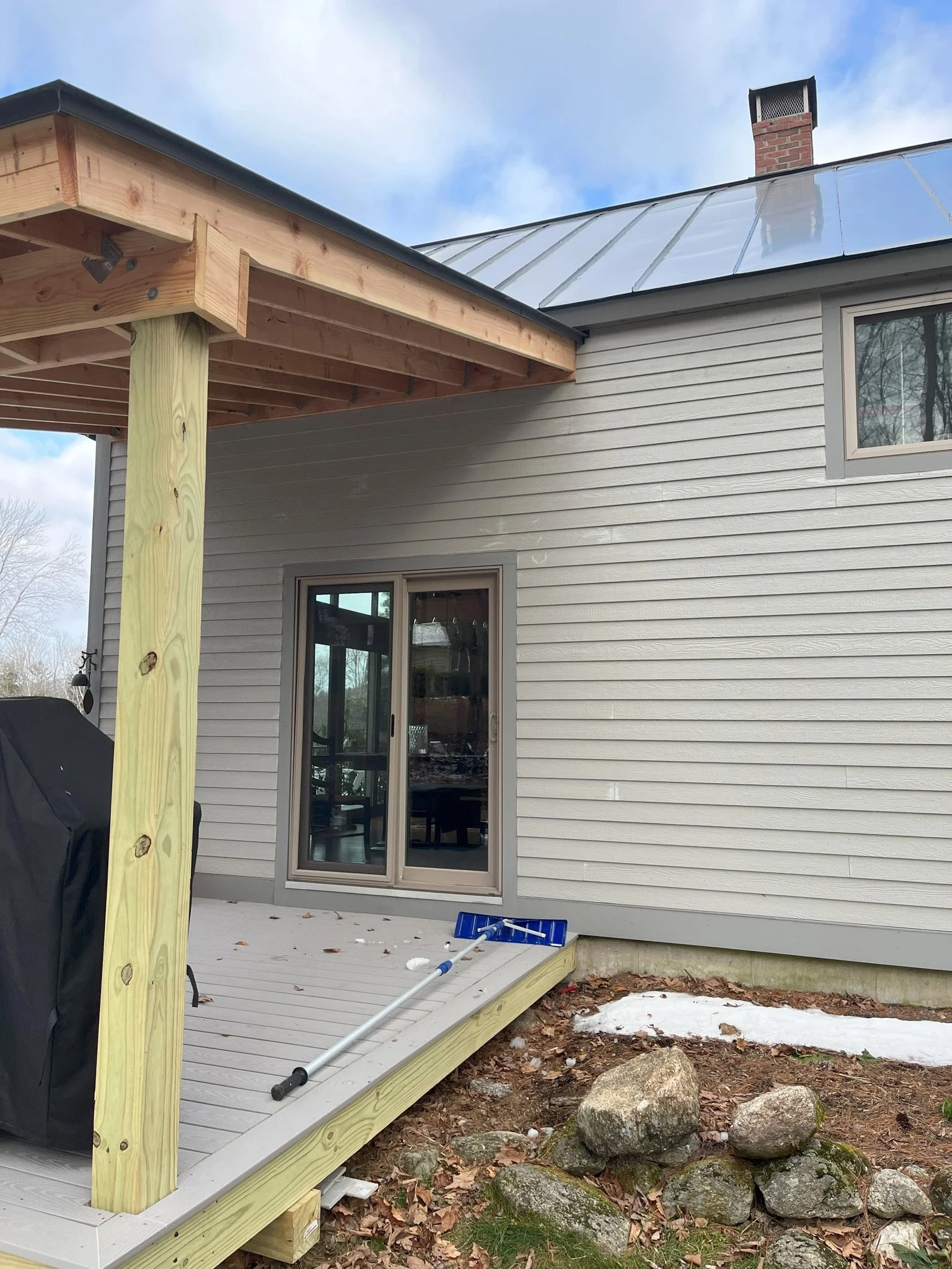 Backyard with wooden deck under construction, sliding glass door, and partially finished siding house with gray exterior, a metal roof, and a brick chimney, with rocks and snow on the ground.