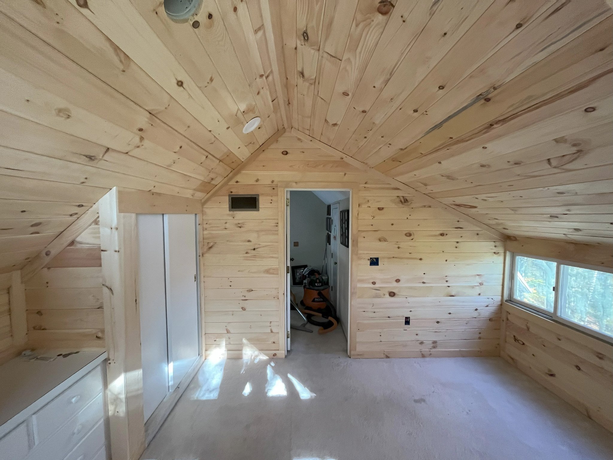 A small, unfinished attic room with wood-paneled walls and ceiling. There is a small window on the right side, a closet with sliding doors on the left, and an open doorway leading to another room. The floor is carpeted, and some items are visible thr