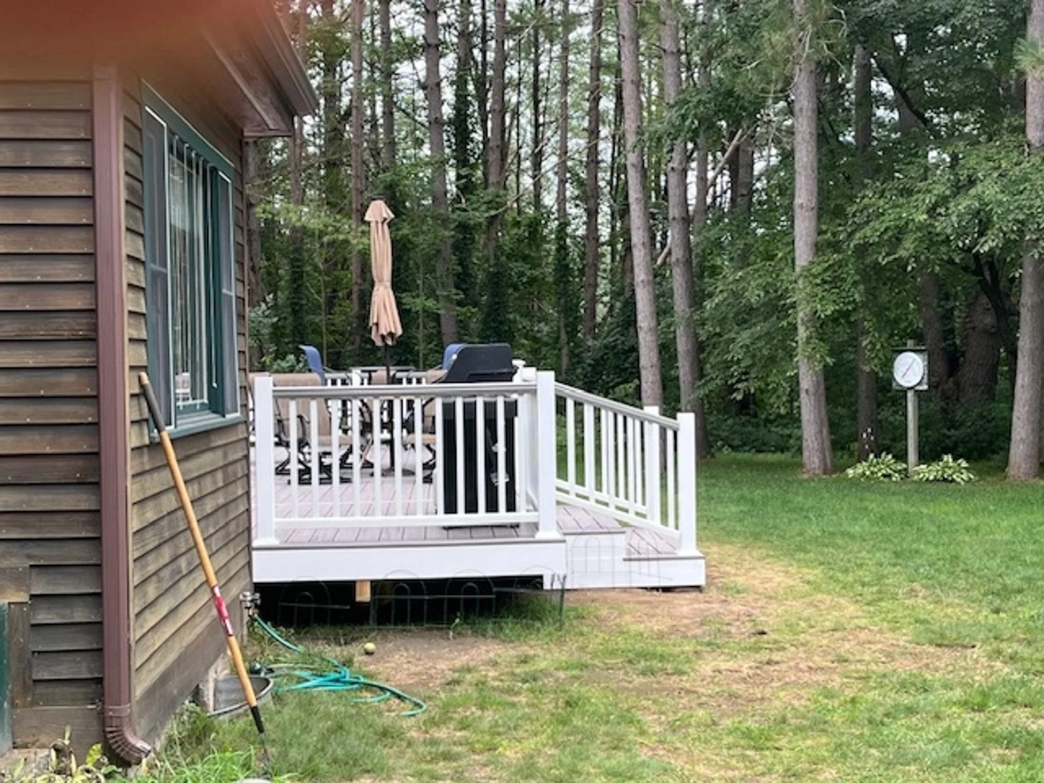 A backyard with a wooden deck attached to a house, located next to a grassy lawn and surrounded by tall trees. The deck has a patio table and chairs, an umbrella, and a black grill. A garden hose and gardening tools are near the house, and there is a