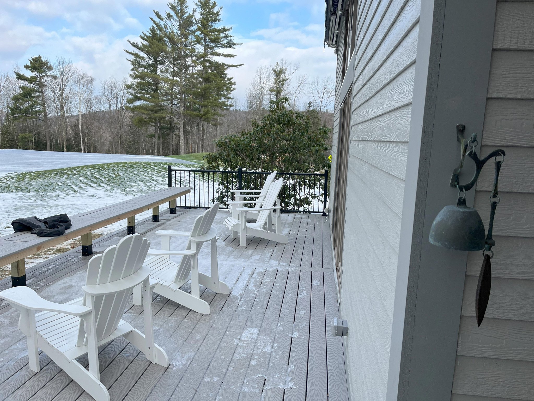 Back porch with four white Adirondack chairs, a black railing, and a wooden bench with a jacket on it, overlooking a snow-dusted lawn with trees in the distance.
