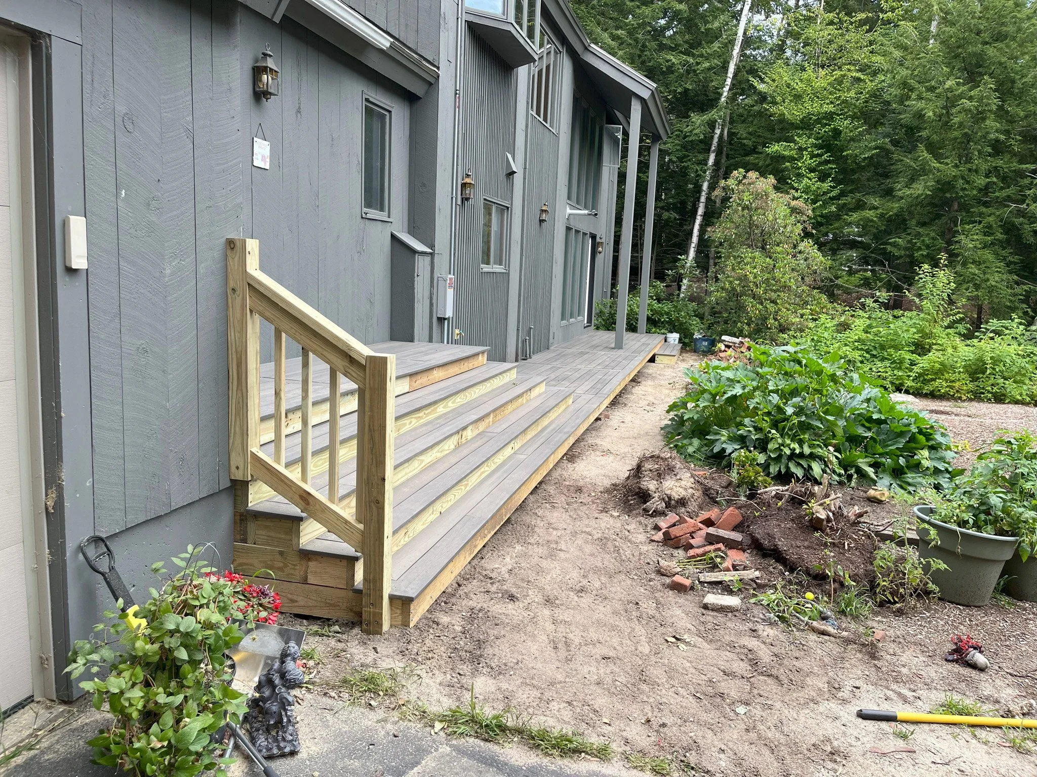 Under construction wooden deck attached to a gray house with a small set of stairs, surrounded by gardening plants, tools, and materials.