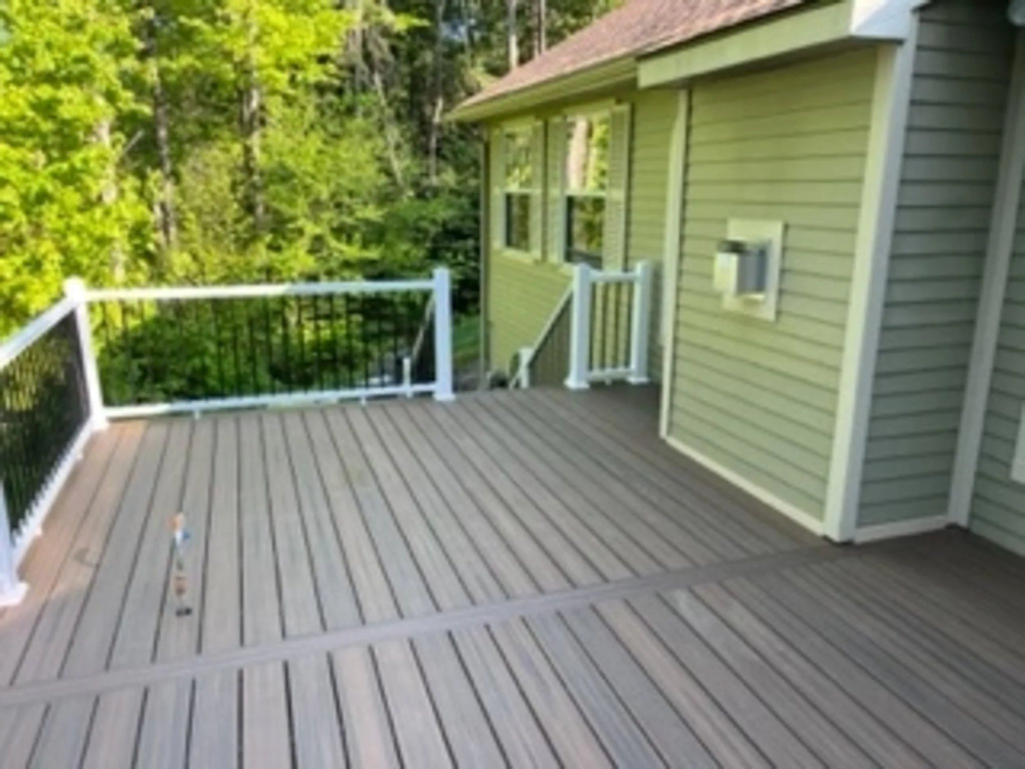 A wooden outdoor deck attached to a house, with green siding, a white railing, and surrounded by trees.