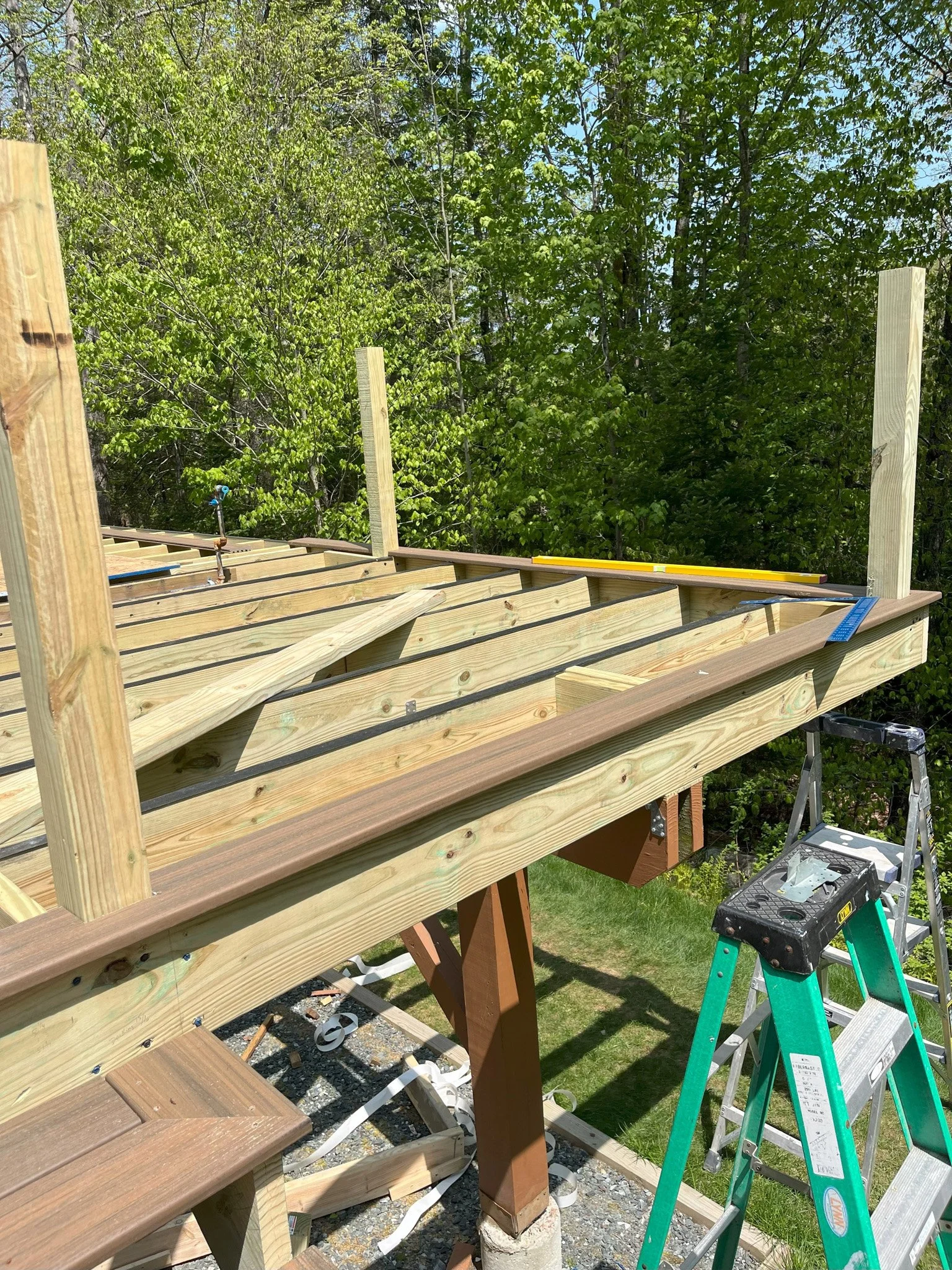 Wooden deck under construction with support beams, a ladder, and construction tools, surrounded by green trees in a backyard.