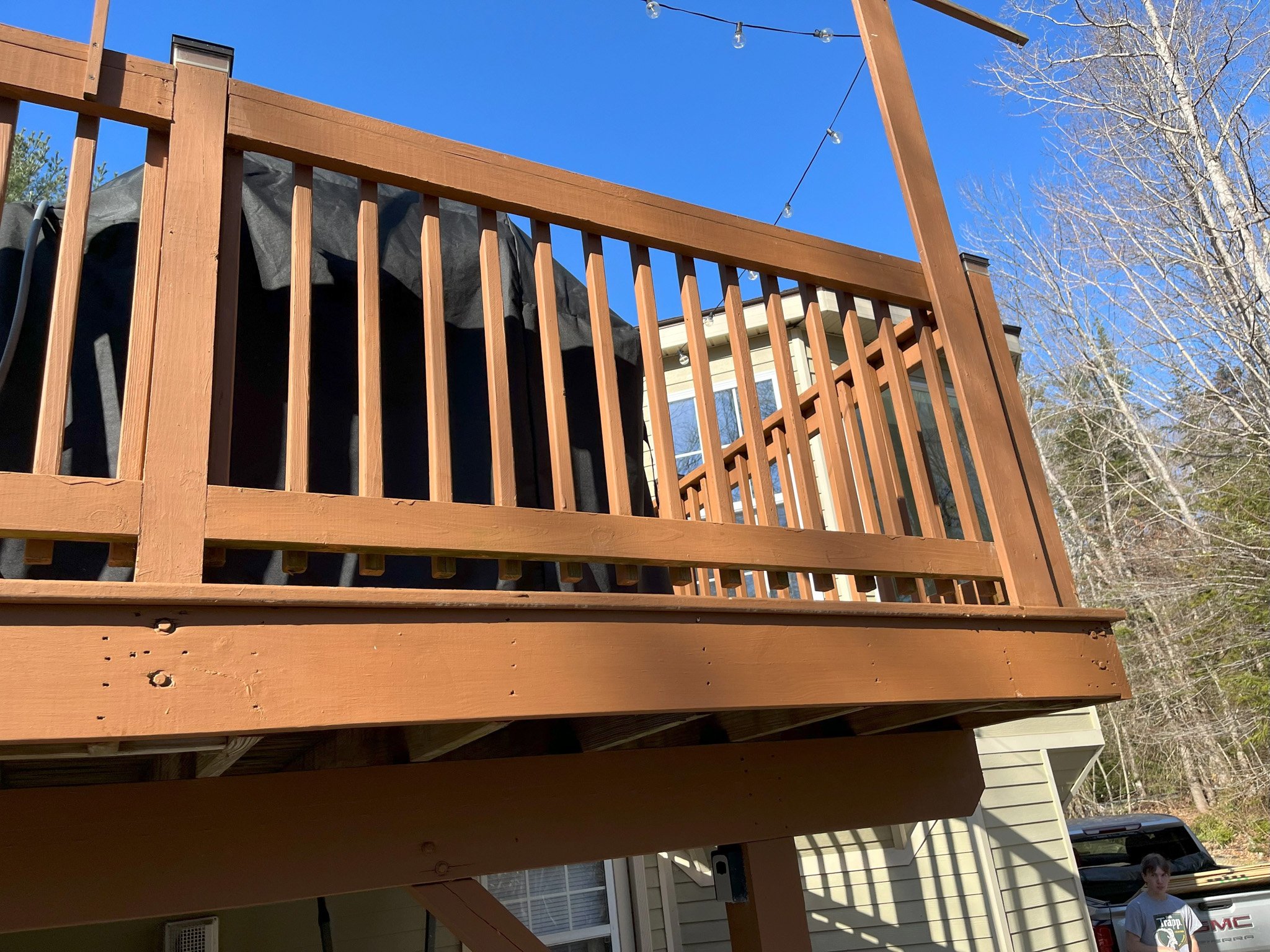 Wooden deck with railing attached to a house, with a person standing nearby, a car parked, trees, clear blue sky, and string lights overhead.