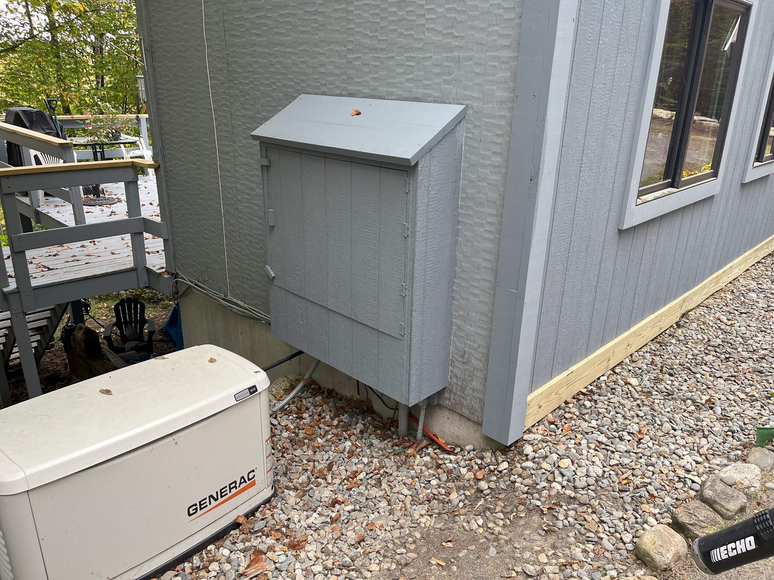 Outdoor view of a gray house with a utility box mounted on the exterior wall and a portable generator on the ground, with a gravel yard, deck, and trees in the background.