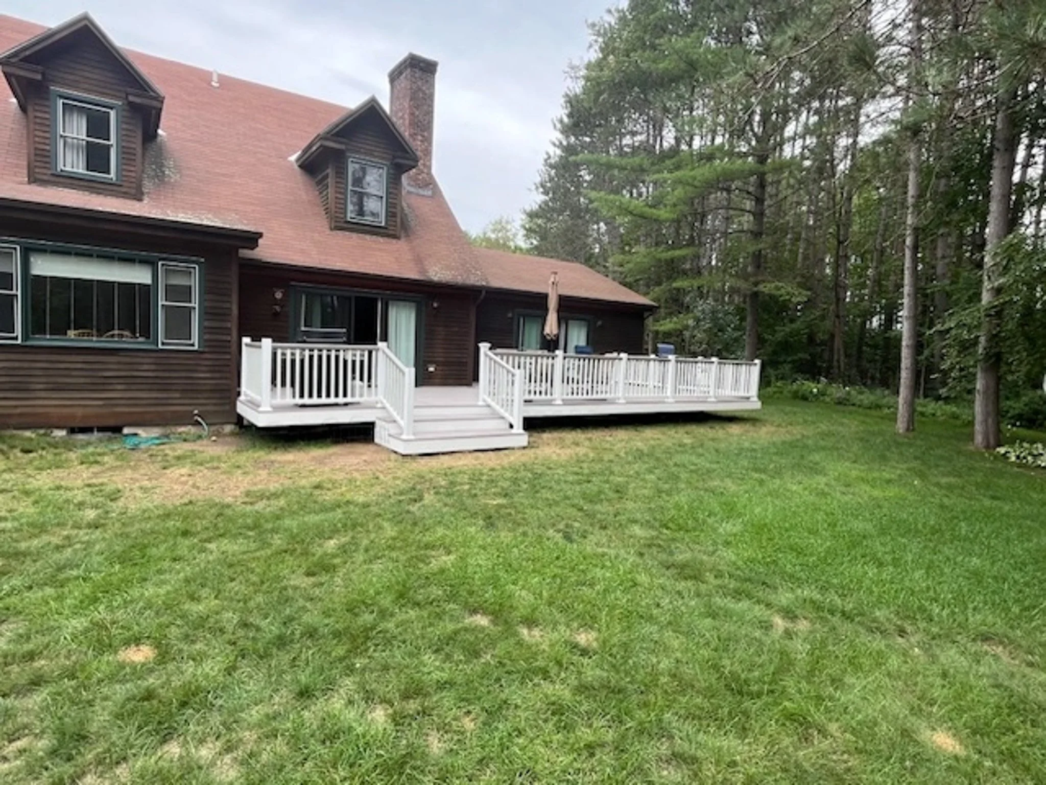 A two-story house with dark wooden siding and a red gabled roof, featuring multiple windows and a white wooden deck in the backyard. The yard has green grass with some patches, and there are trees in the background.