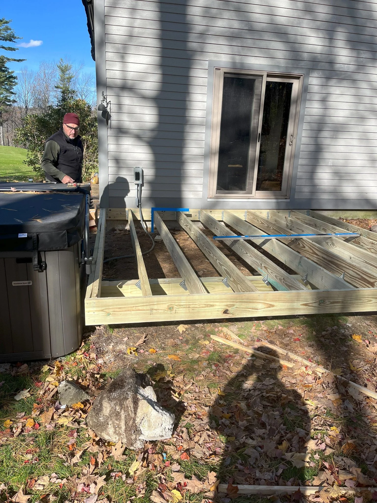 A man wearing a beanie hat, glasses, and a black vest stands next to a partially built wooden deck frame attached to a house with gray siding. There is a hot tub on the left side and visible trees and grass in the background. The photo is taken outdo